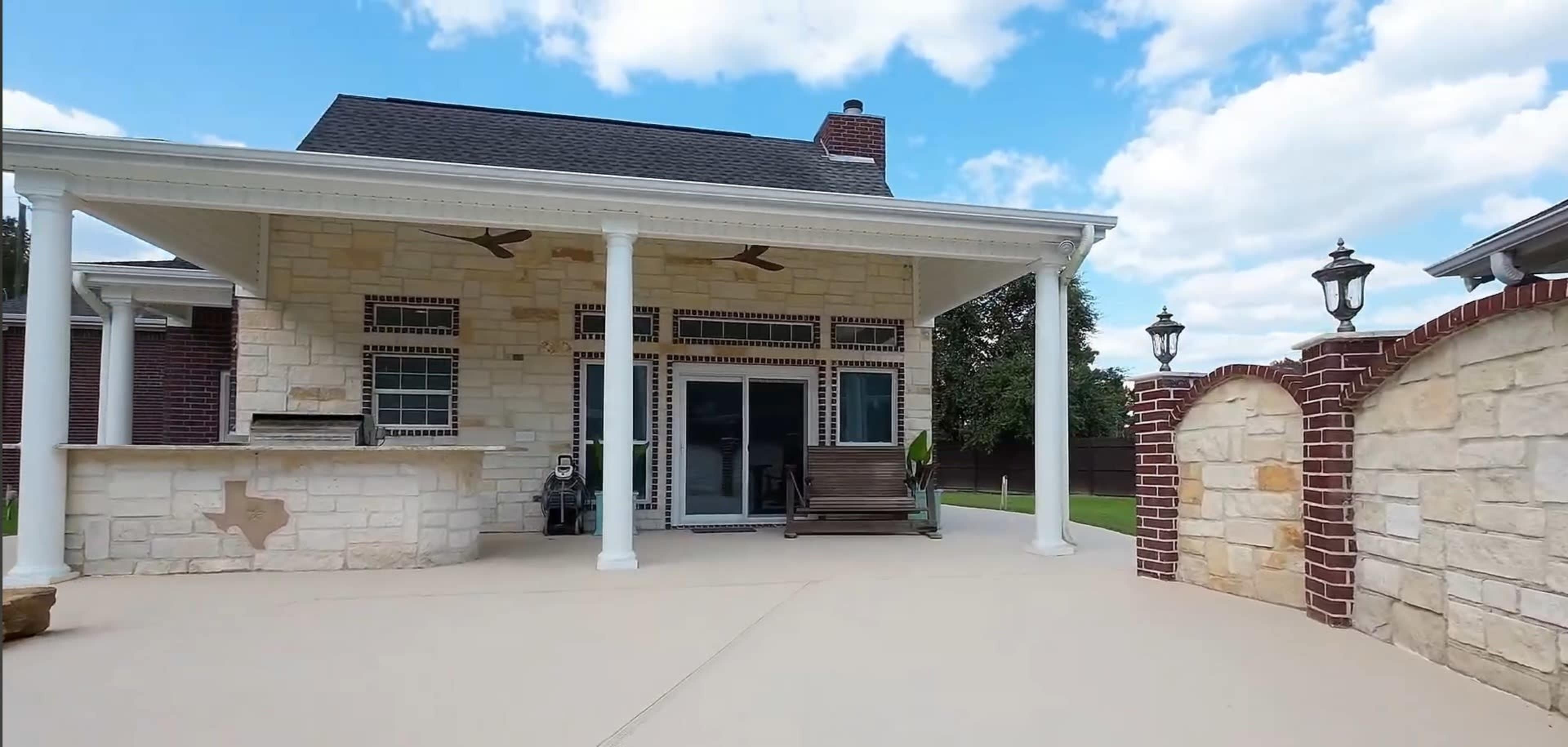 The image shows a spacious outdoor patio area featuring a stone house with a covered porch, a wooden bench, and decorative lampposts against a backdrop of blue sky and fluffy clouds.