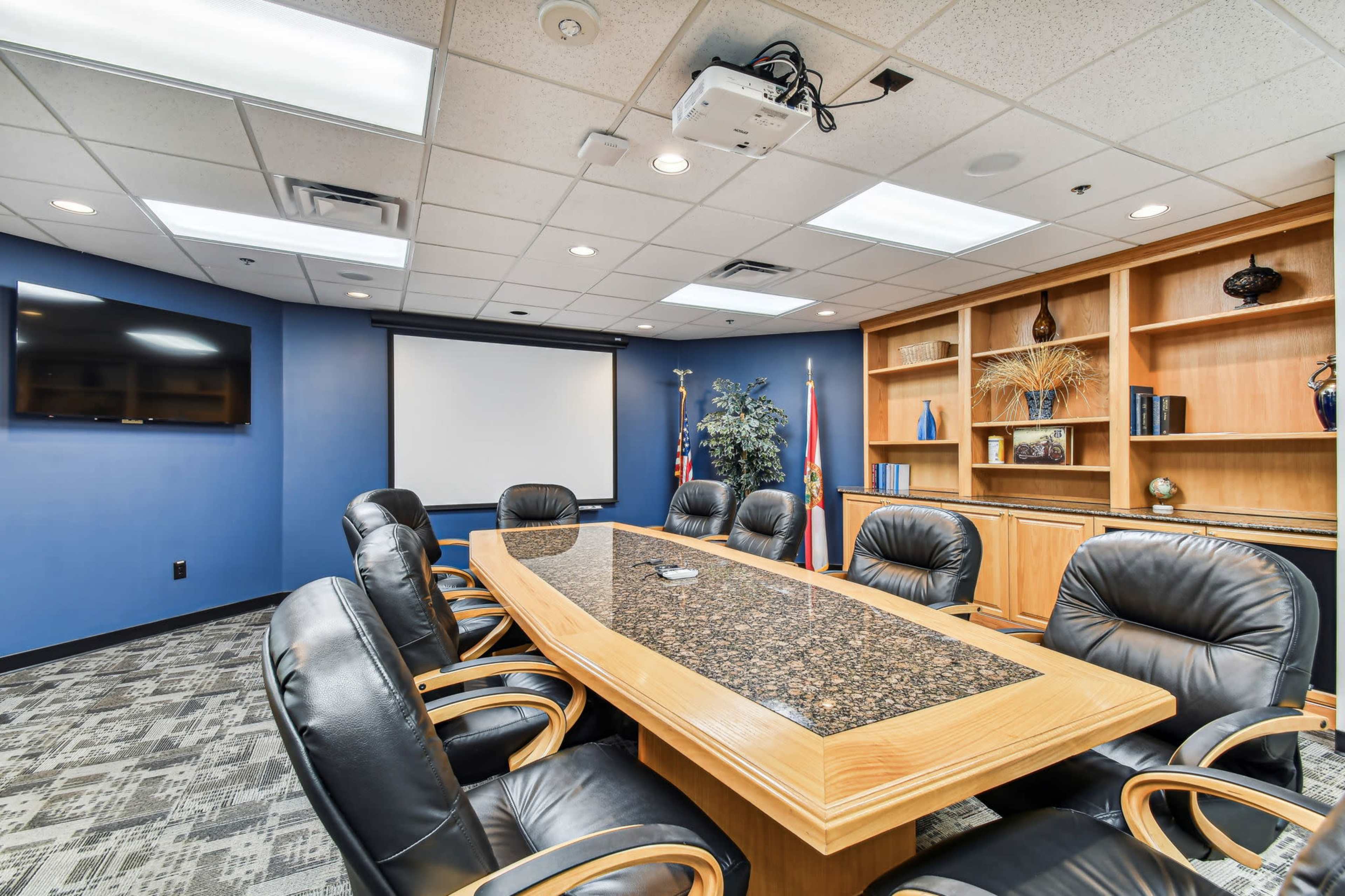 The image shows a modern conference room with a large wooden table surrounded by black leather chairs, a projector, and a wall-mounted screen.
