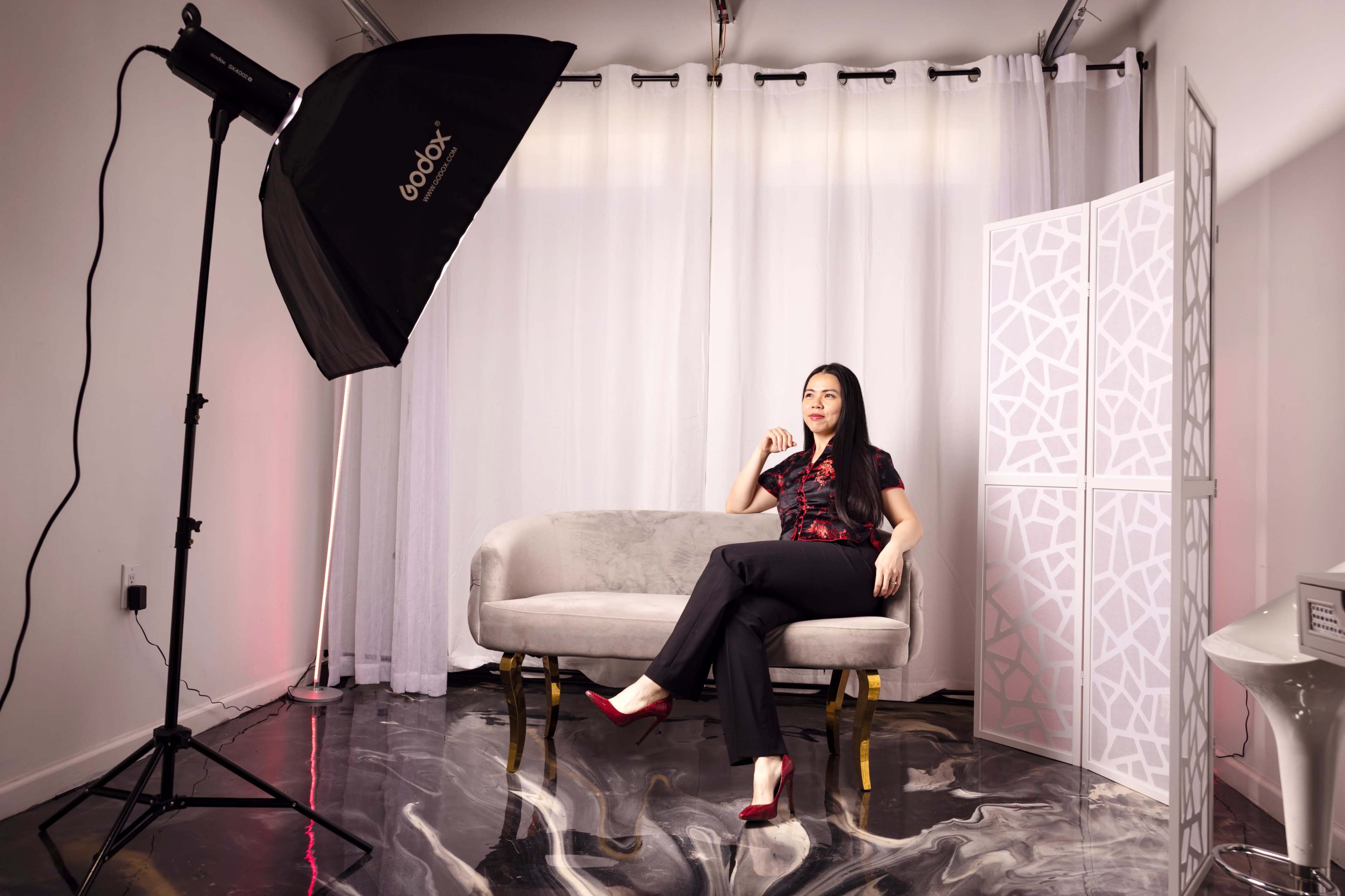 A woman in a black and red outfit sits on a gray loveseat, with a large lighting setup and a folding screen in the background.