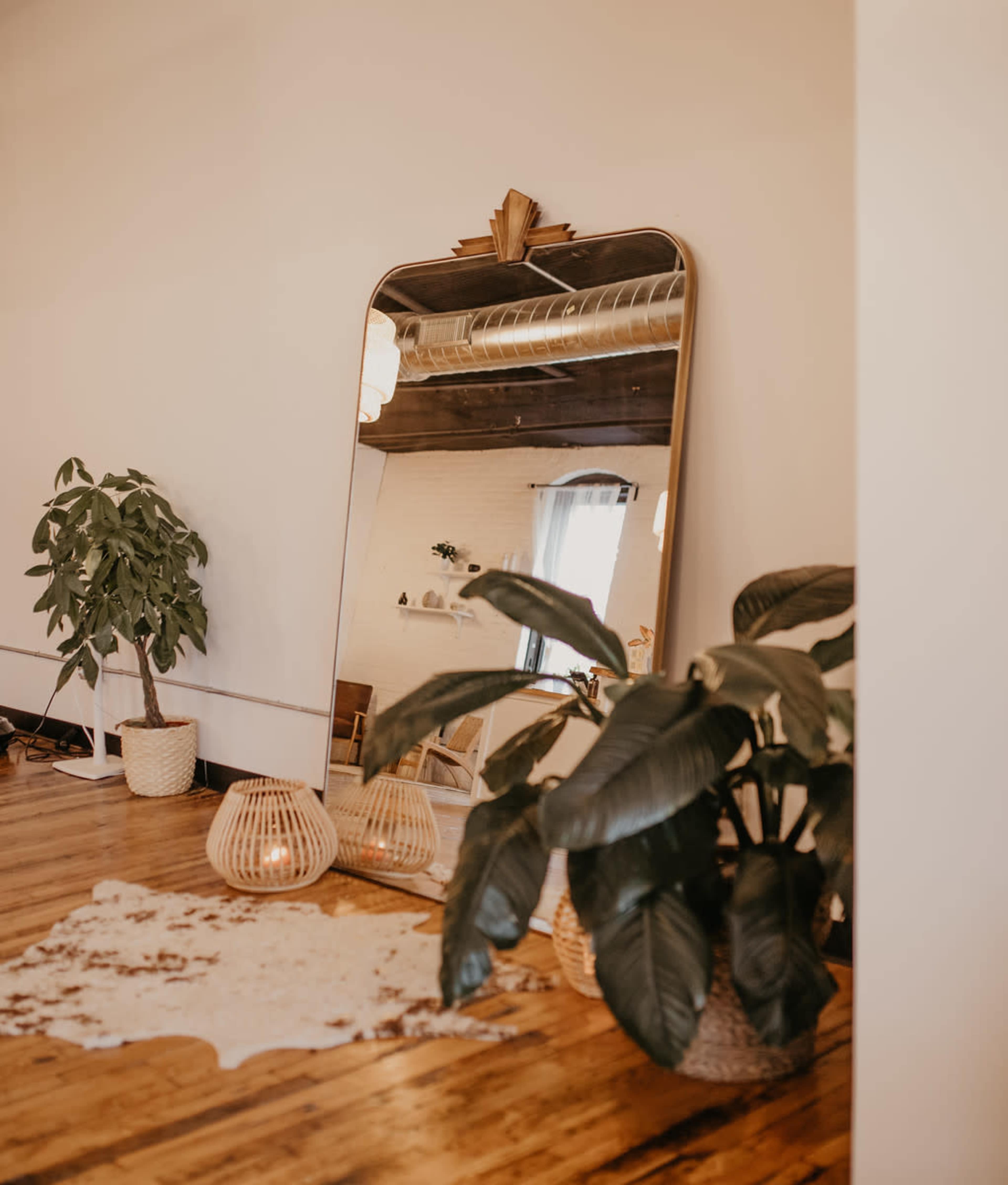 A large arched mirror reflects a room with a plant, wicker baskets, and a cowhide rug on a wooden floor.