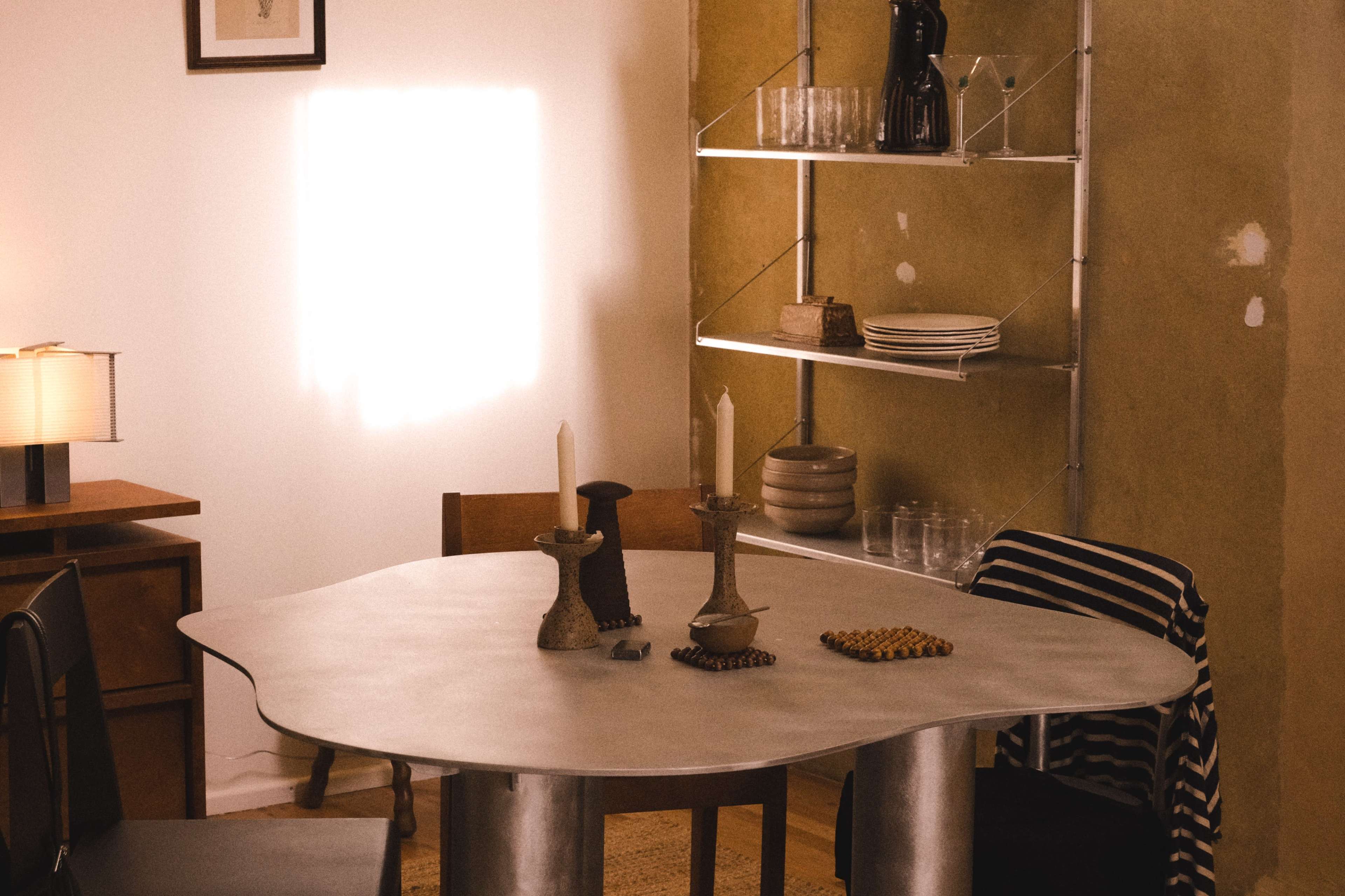 A modern dining table with a unique shape is set in a minimalist room, featuring two candlesticks and shelves filled with glassware and plates.
