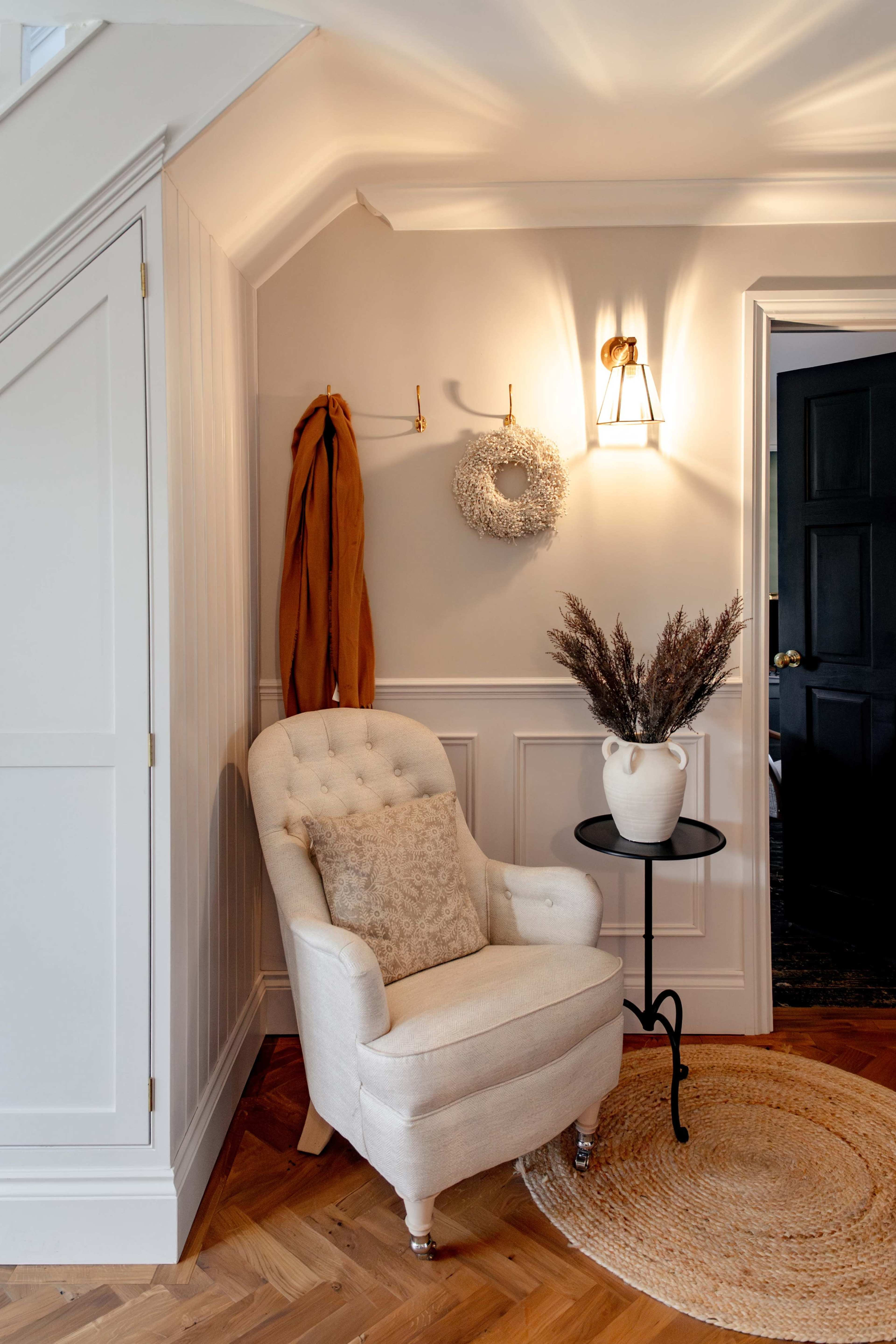 A cozy corner features a white upholstered armchair with a decorative pillow, a small black side table, and a potted plant against a light gray wall with hooks and a wreath.