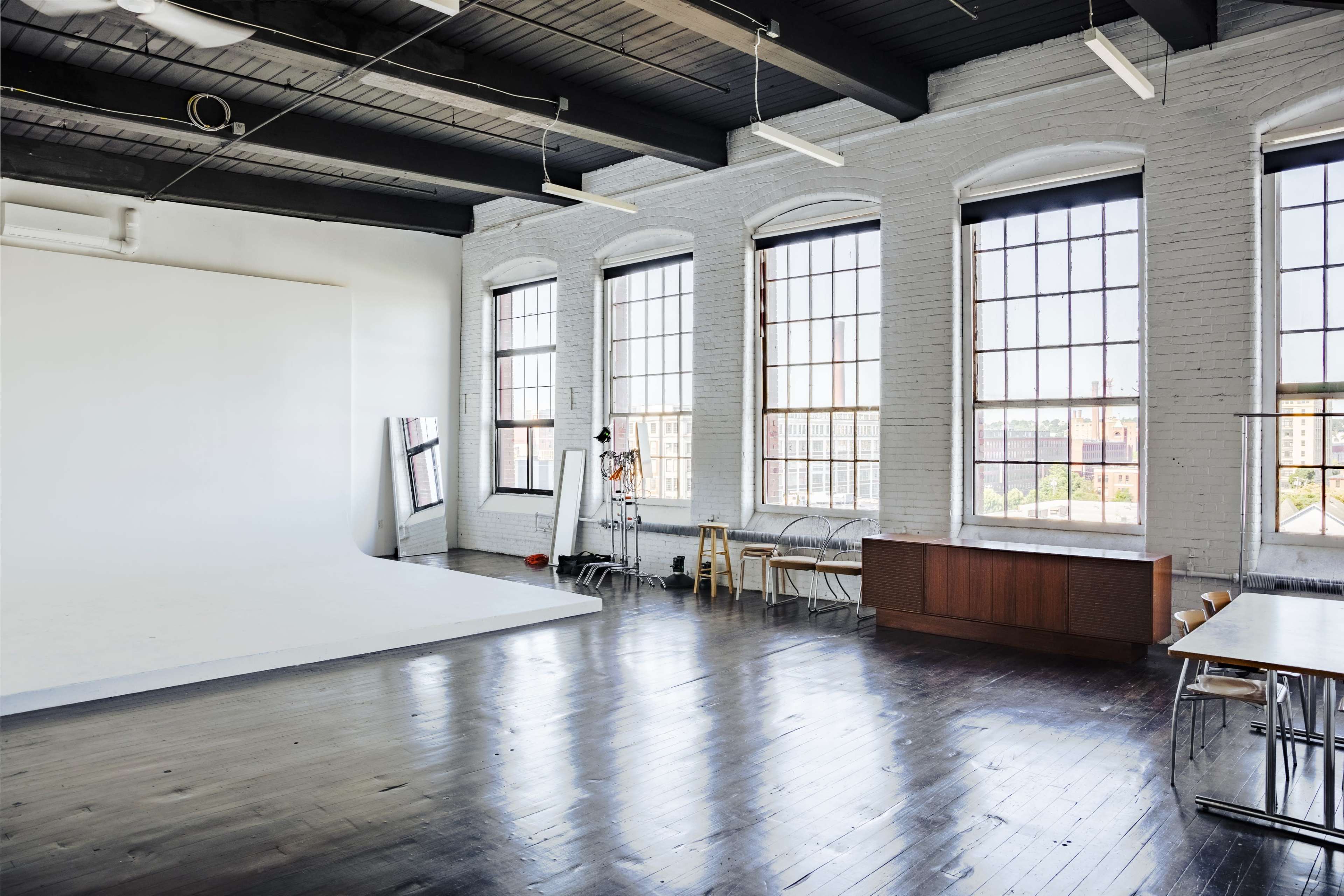 A spacious photography studio with large windows, a plain white backdrop, and several pieces of furniture arranged along the walls.