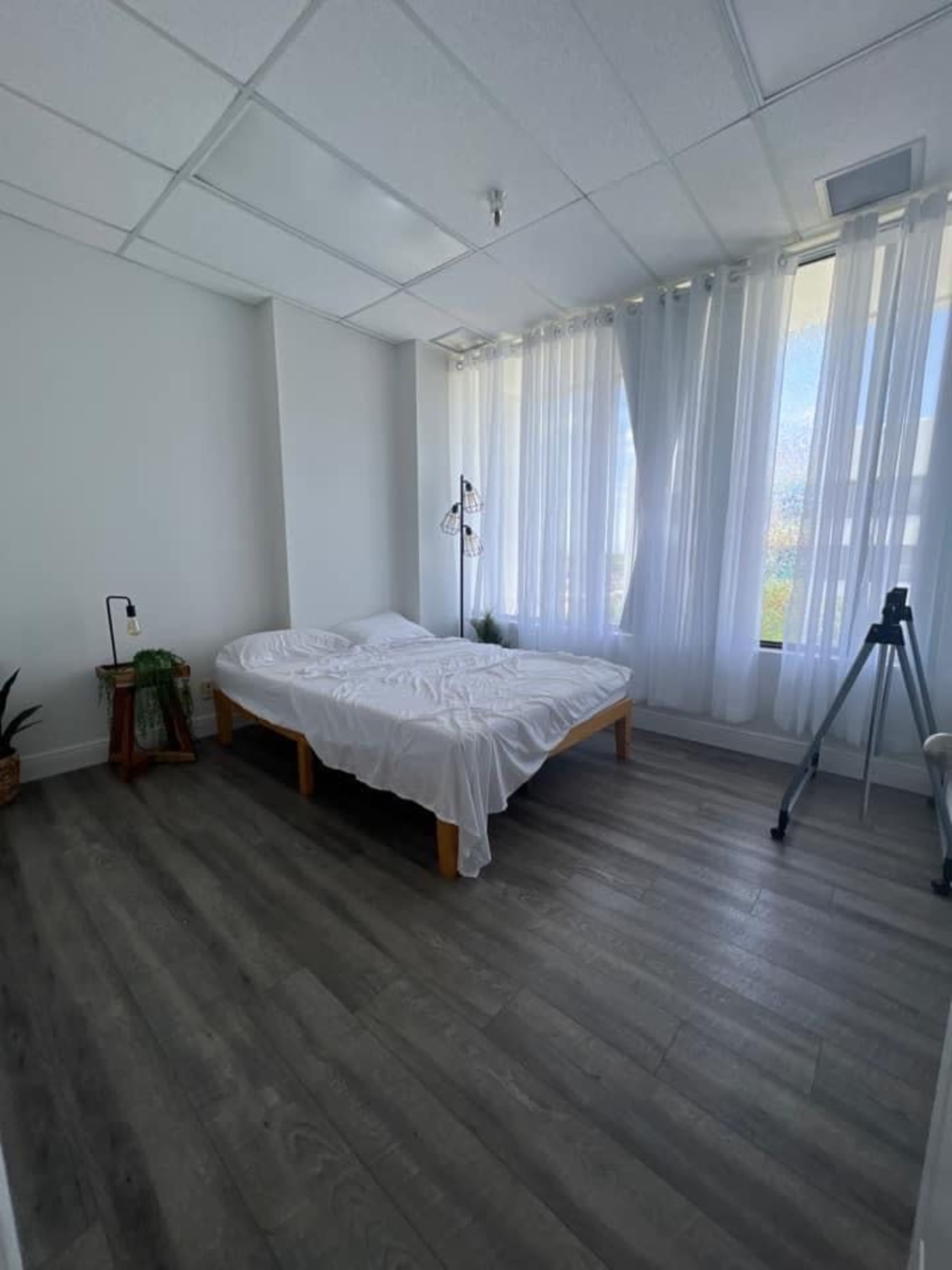 A minimalist bedroom featuring a bed with white bedding, large windows covered by sheer curtains, and wooden flooring.