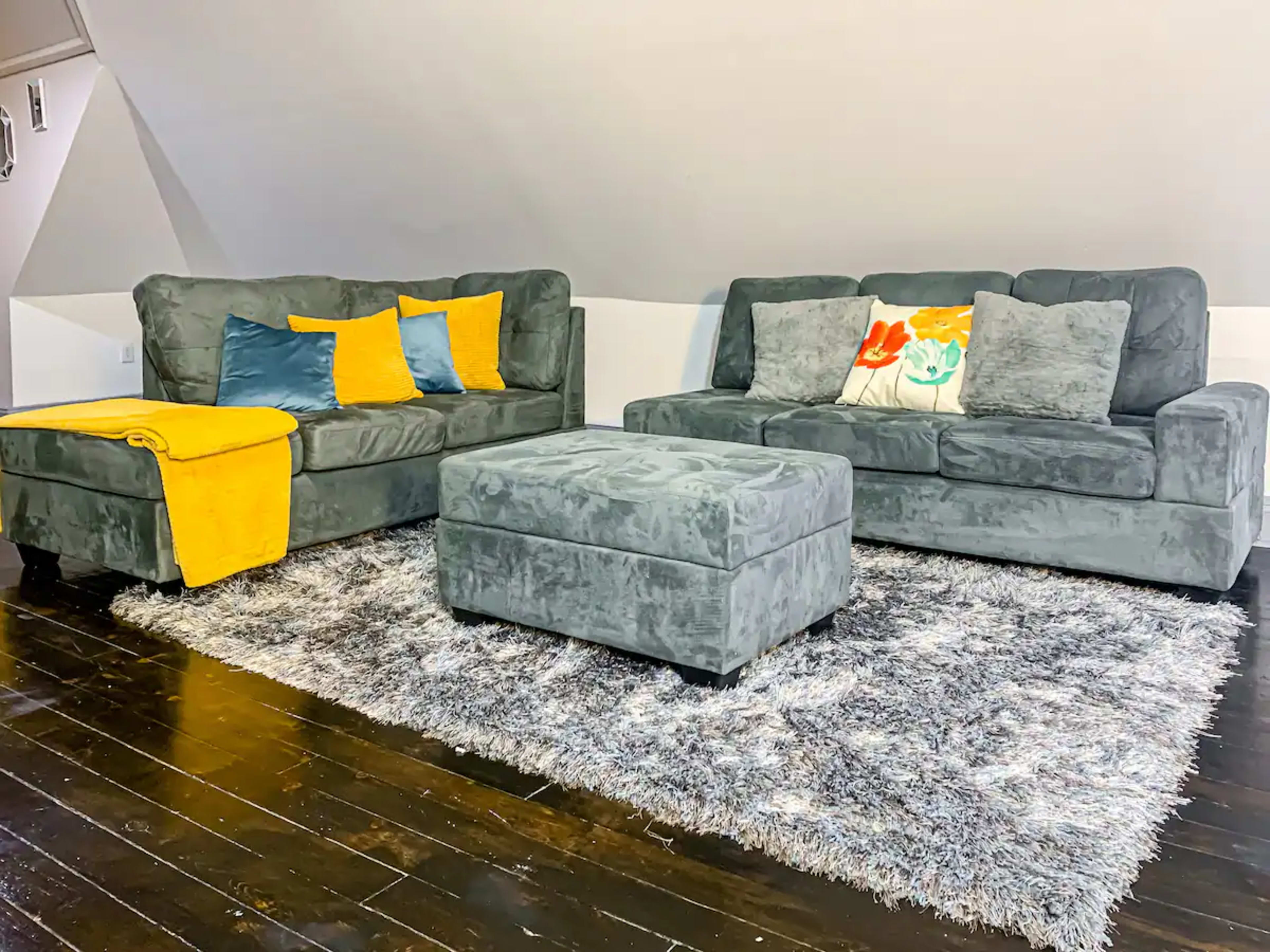A gray living room setup featuring a loveseat and a sofa with colorful pillows, accompanied by a matching ottoman and a yellow throw blanket on one end, all placed on a plush area rug over dark wooden flooring.