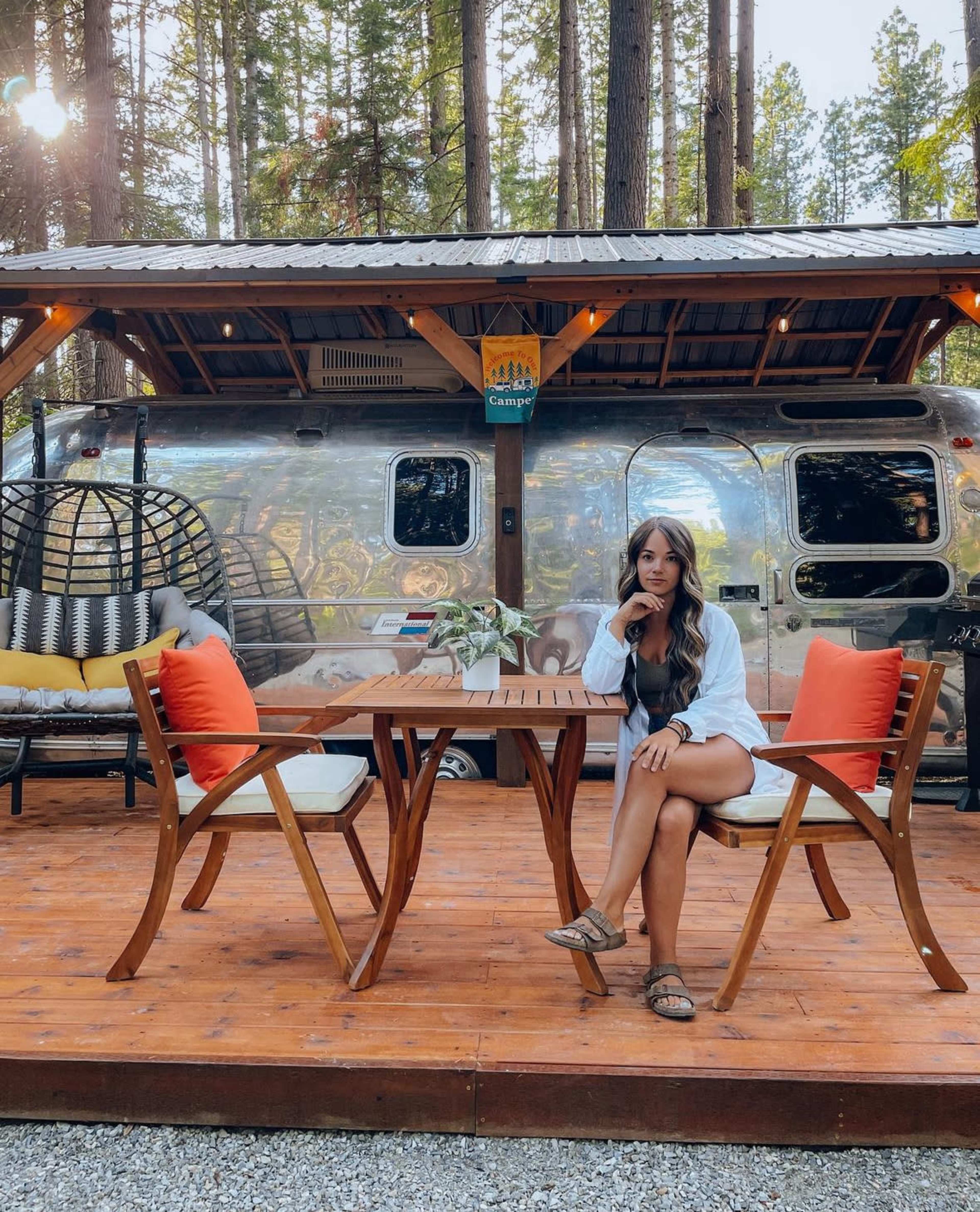 A woman sits at a wooden table on a patio with chairs, facing a shiny Airstream trailer surrounded by tall trees.