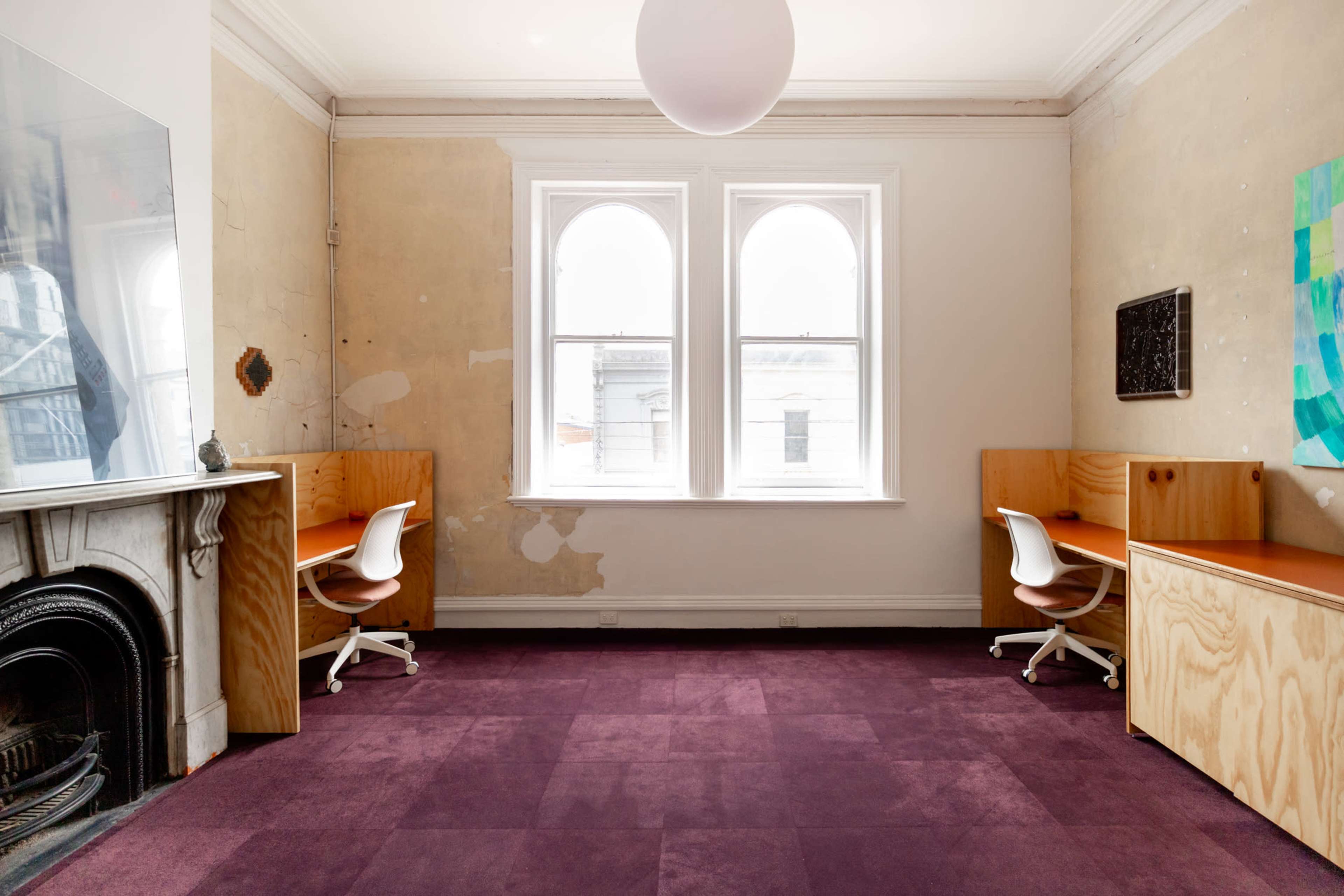 The image shows a spacious room with two wooden desks, white chairs, and purple carpeting, featuring windows on one wall and partially exposed walls.