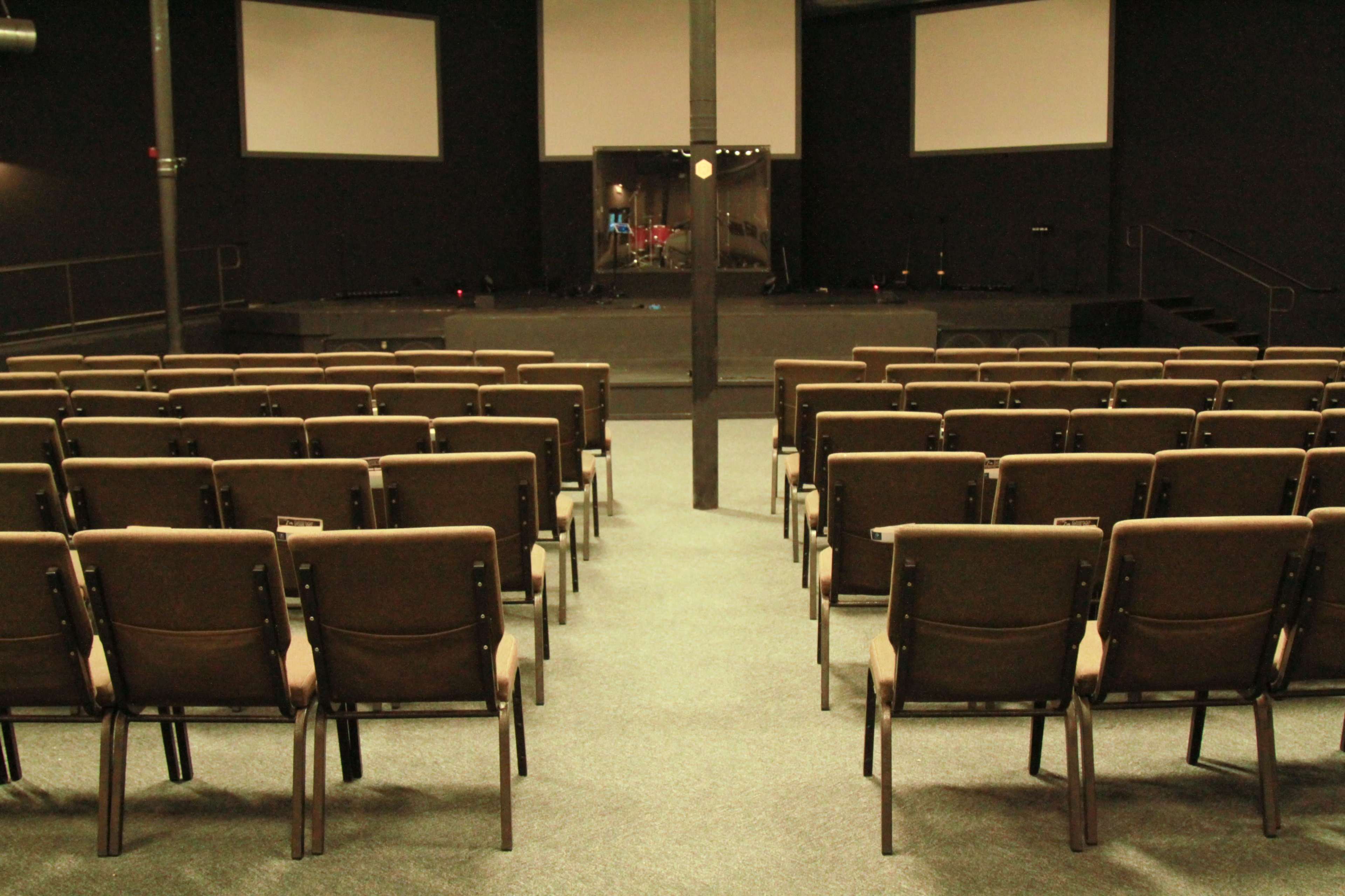The image shows a spacious indoor venue with rows of brown chairs facing a stage that is equipped with lighting and musical instruments.
