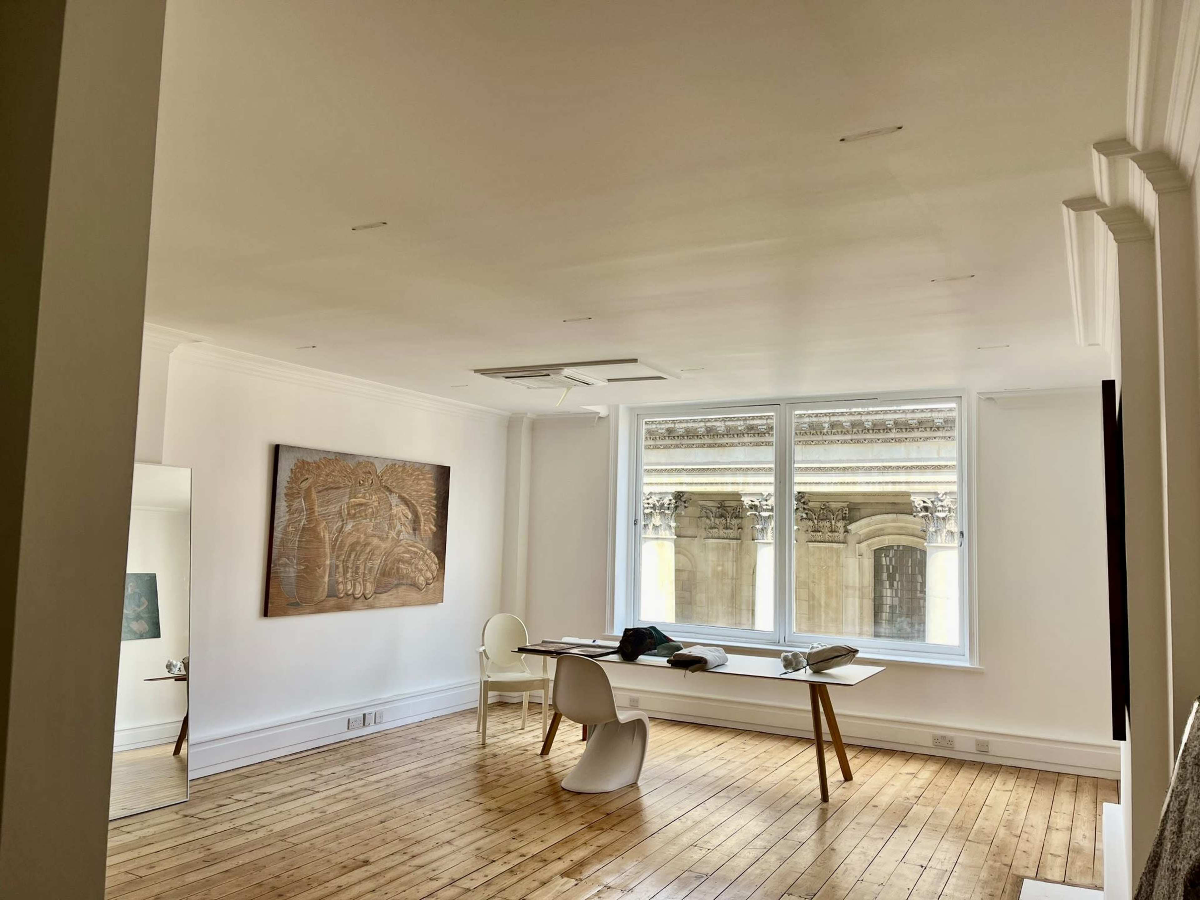 A modern, minimalist room featuring a wooden floor, a large window overlooking a building facade, a table with a few items on it, and a wall with an artistic panel.