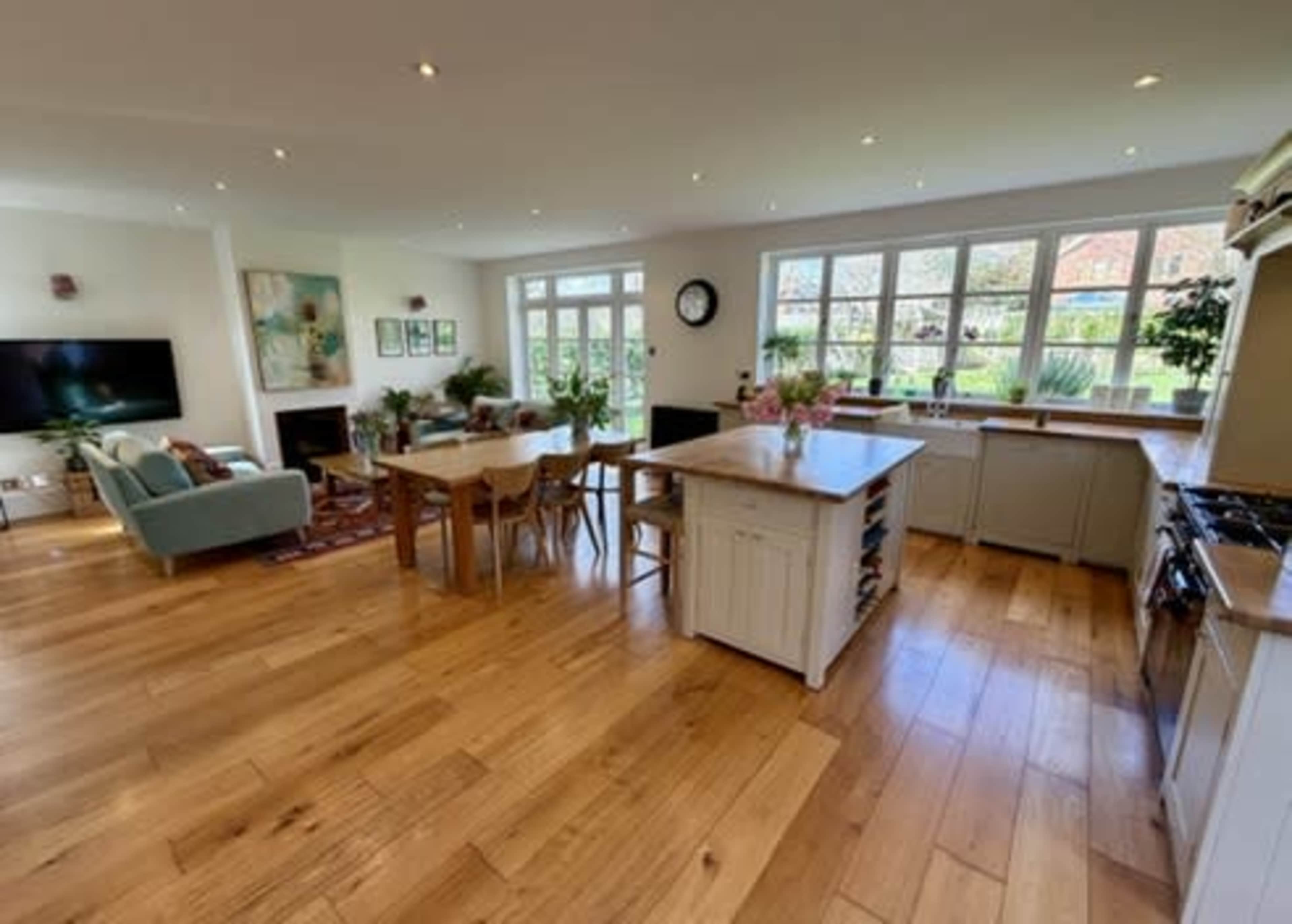 The image shows a spacious, open-concept living and dining area with wooden flooring, large windows, and modern furniture.
