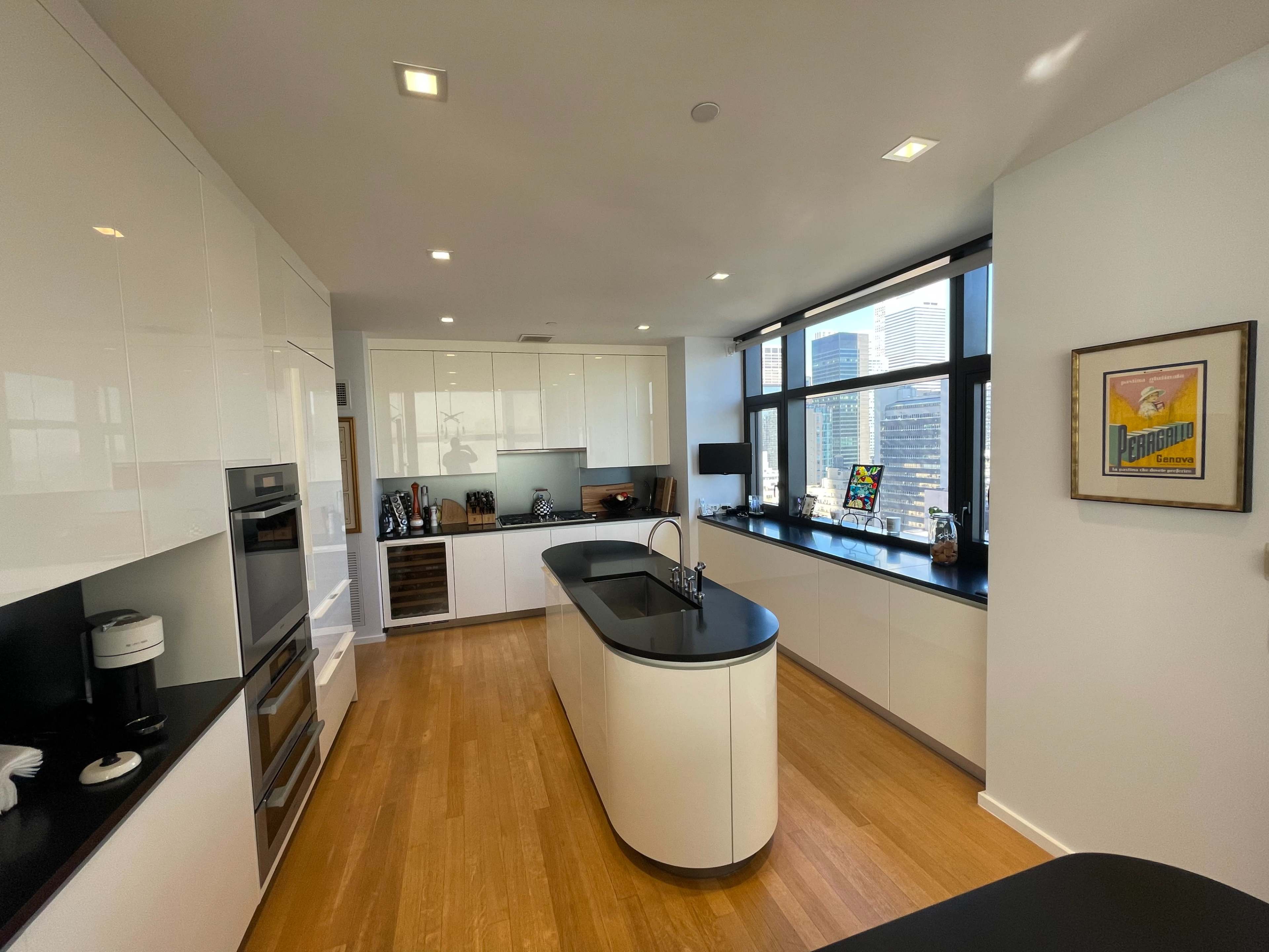The kitchen features modern white cabinetry, a central island with a black countertop, and large windows showcasing a cityscape.