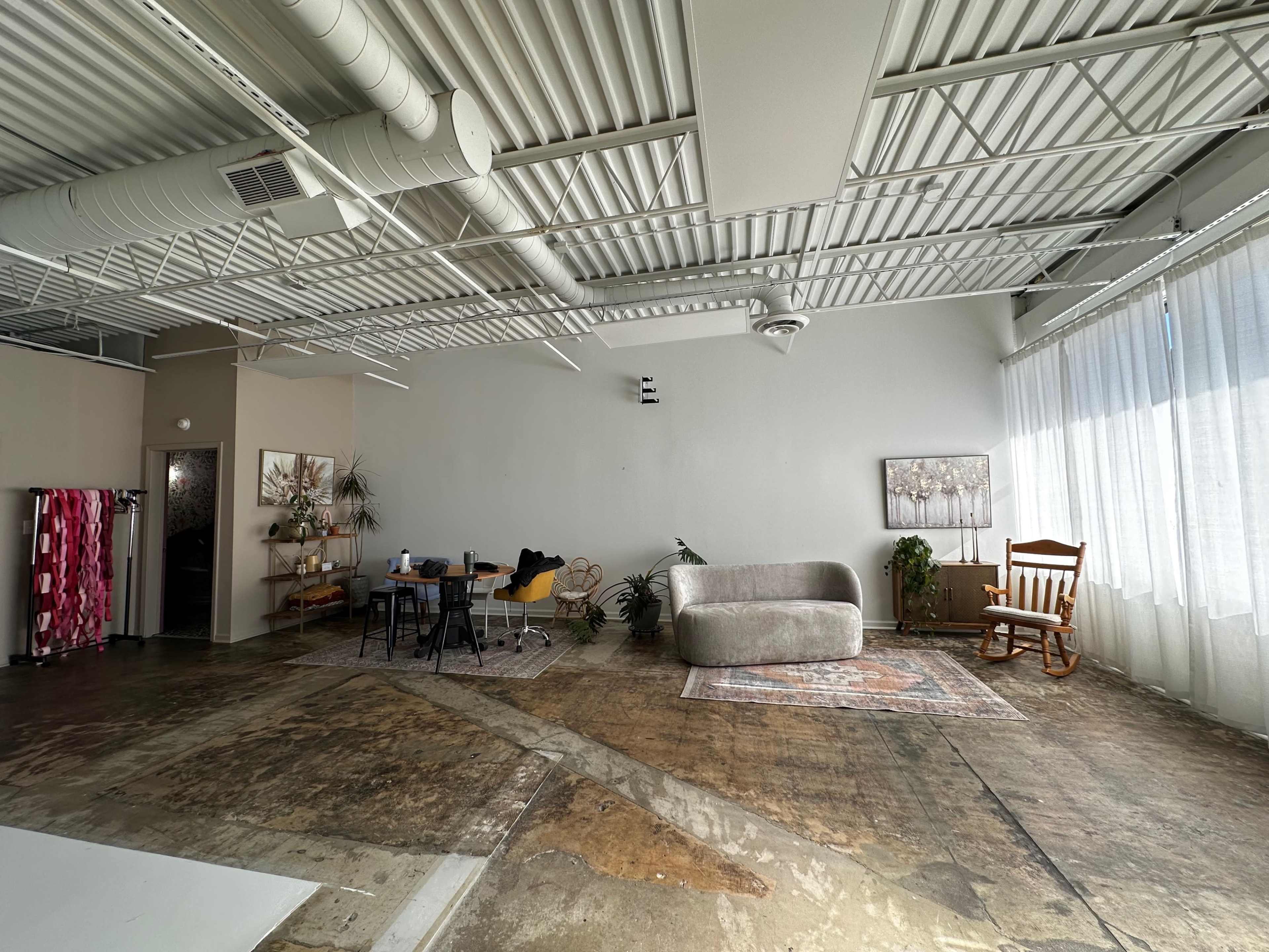 A spacious, industrial-style room with exposed ceilings, a large window, a couch, a dining table with chairs, and a rocking chair, all set against a concrete floor.