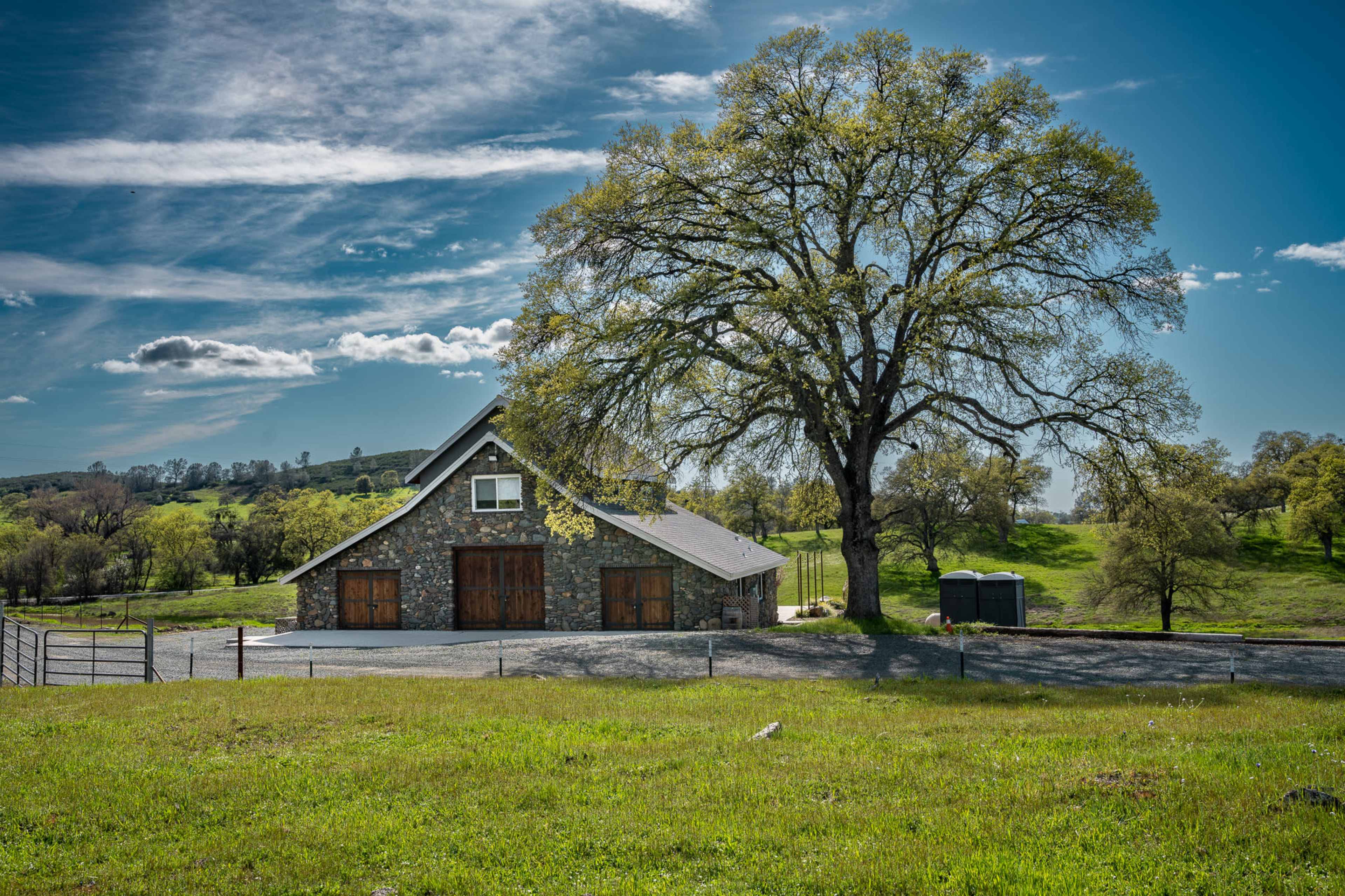 A stone barn with wooden doors stands beside a large tree on a grassy field under a blue sky with scattered clouds.