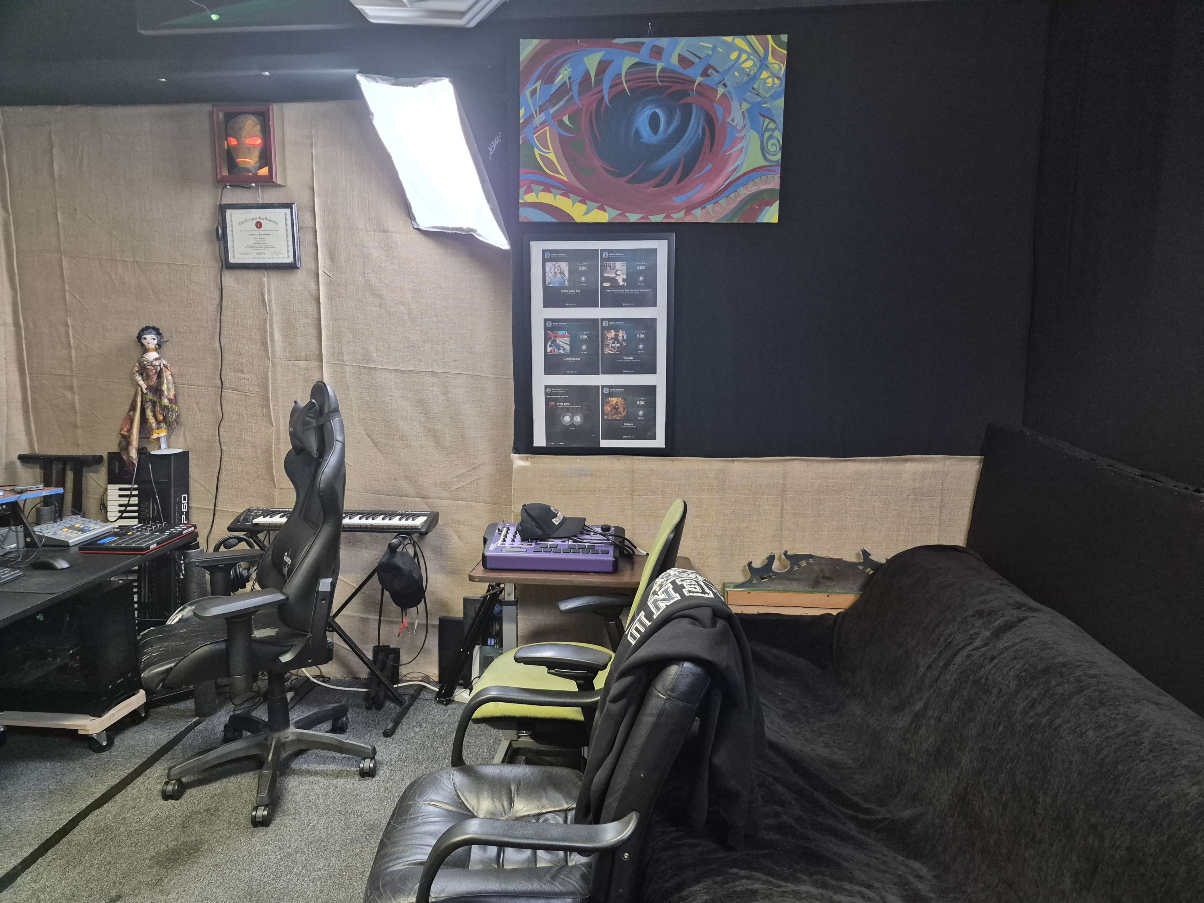 The image depicts a music studio or creative workspace featuring a keyboard, a chair, a couch, a framed wall with artwork and awards, and a figure standing next to the desk.