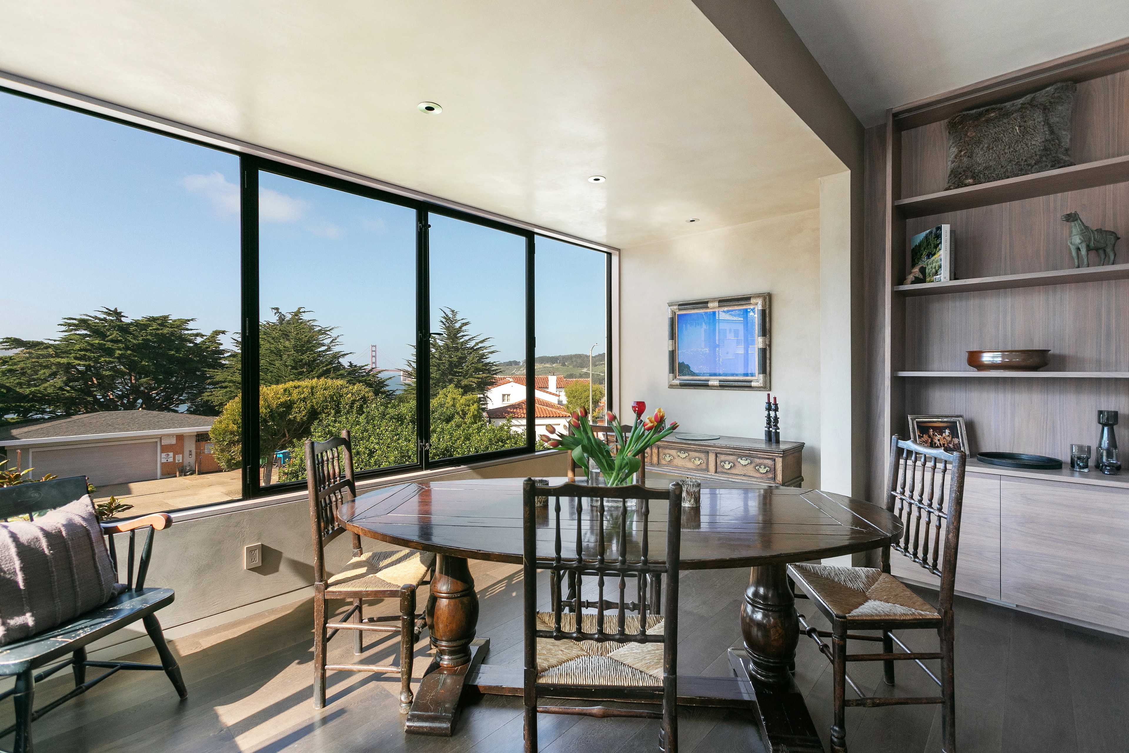A dining area features a round wooden table surrounded by six chairs, with large windows offering a view of trees and homes outside.