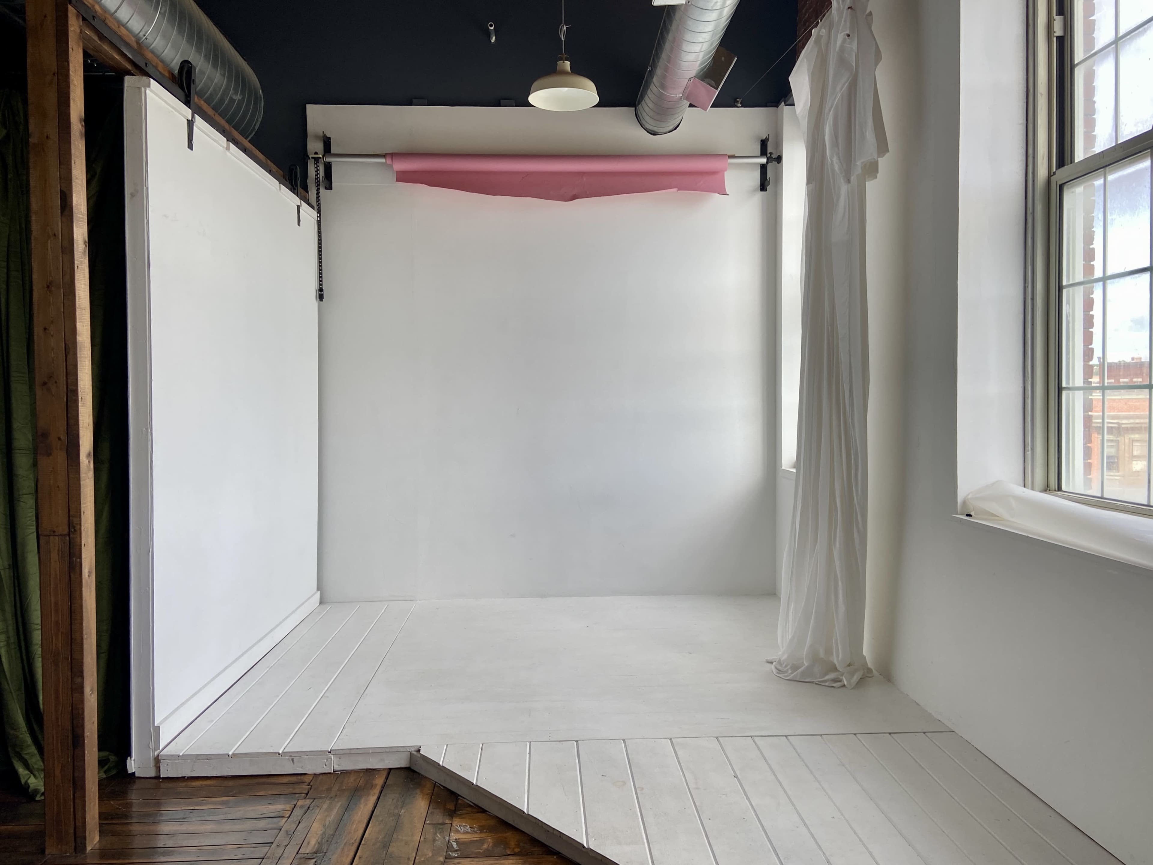 A minimalistic photography studio with a white backdrop, wooden flooring, and a pink paper roll suspended from the ceiling.