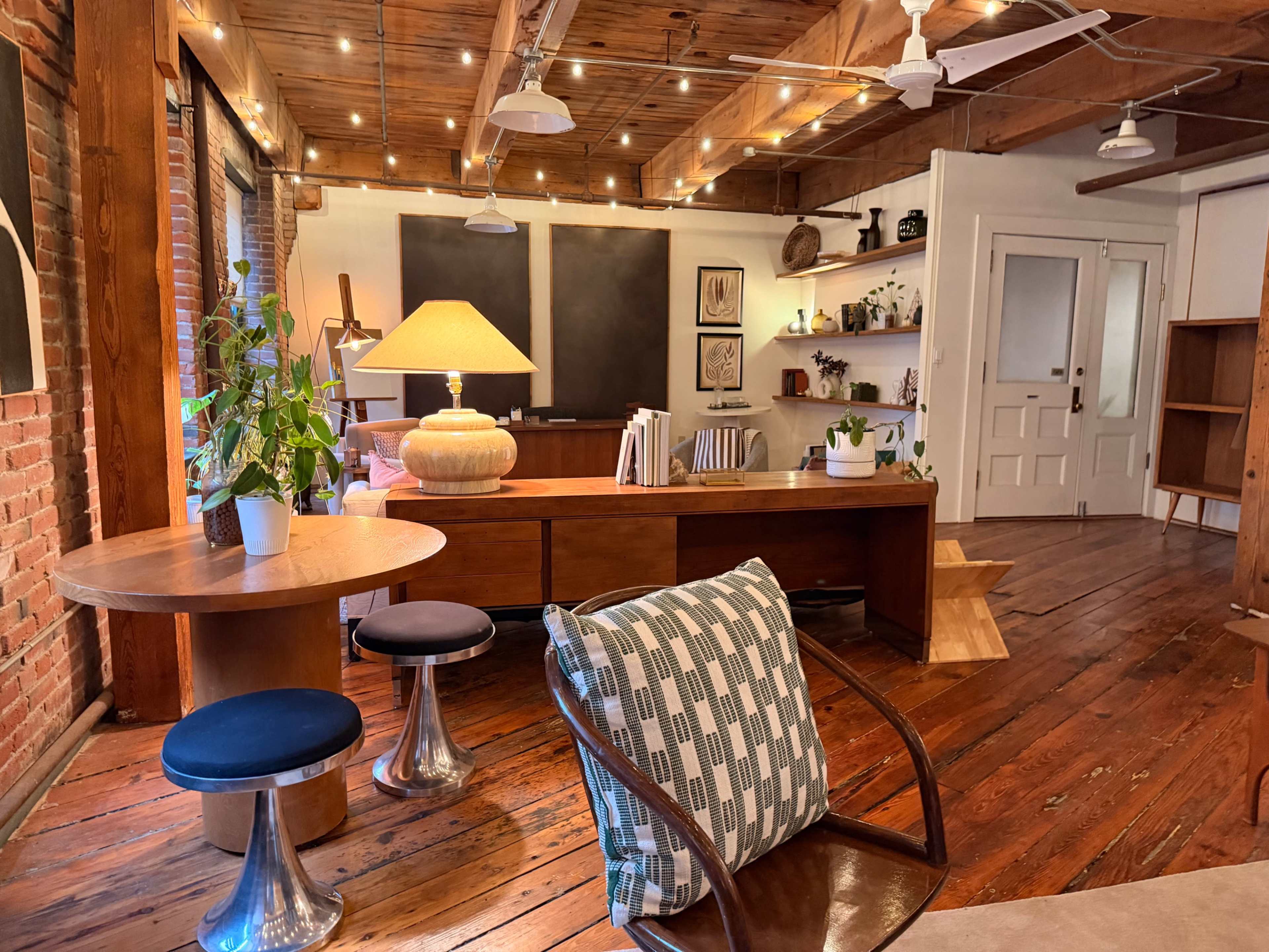 The image shows a well-lit room featuring wooden flooring, exposed brick walls, and modern furniture, including a round table and a cozy lounge chair.