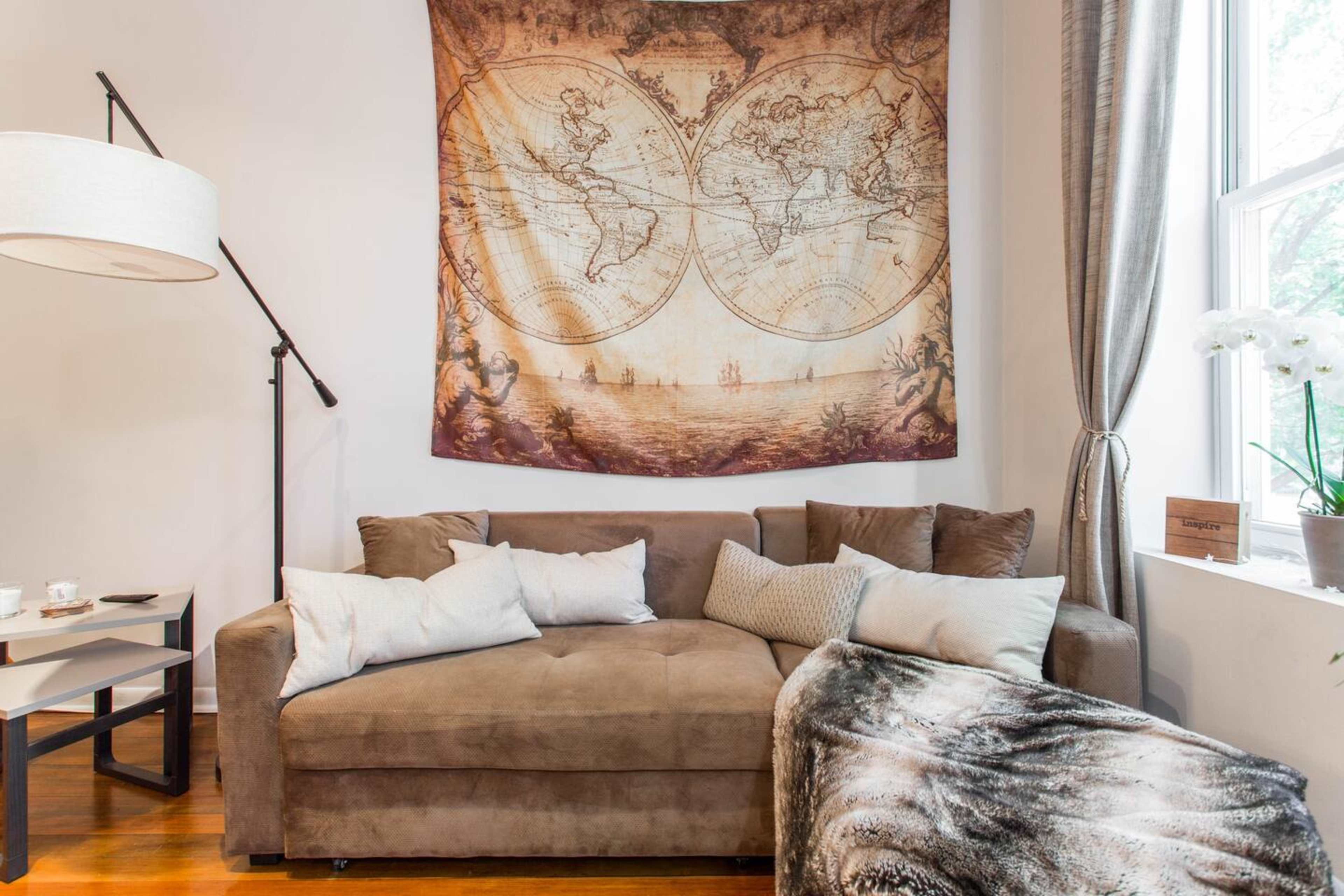 A brown sofa with several pillows is positioned beneath a large vintage world map tapestry, next to a window with sheer curtains.