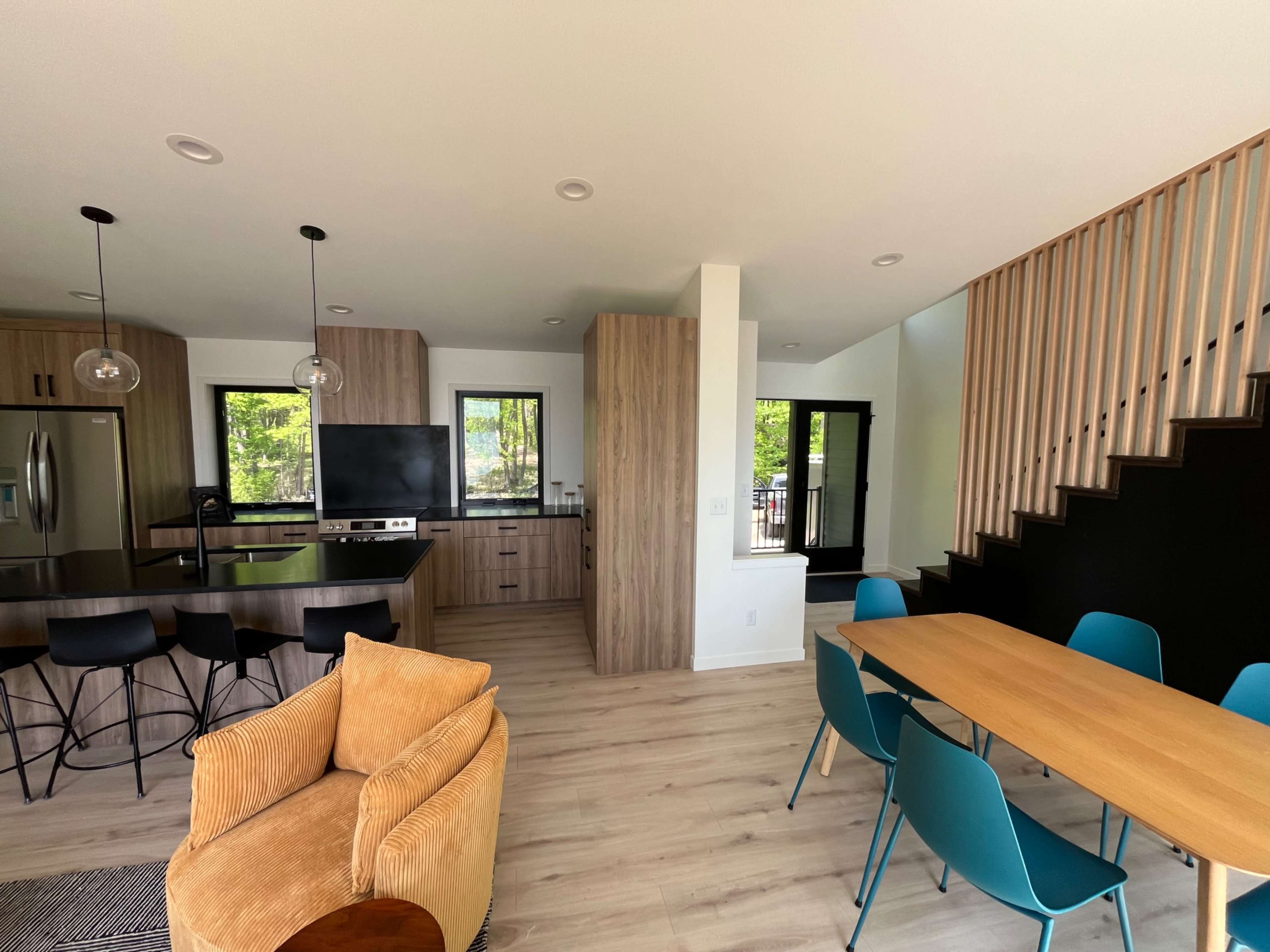 The image shows a modern open-concept living space featuring a kitchen with an island, a dining table with teal chairs, and a staircase leading to another level.