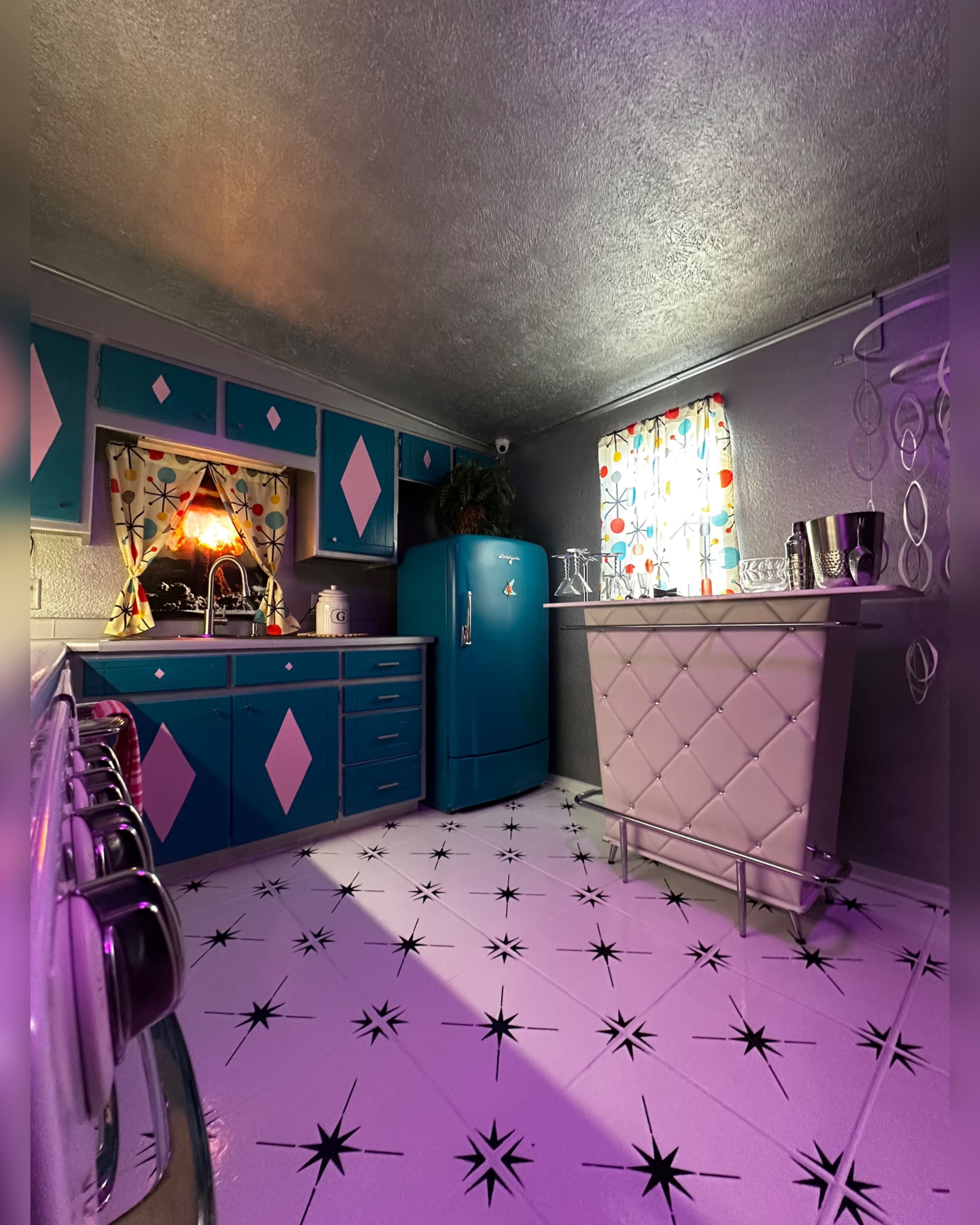 The image shows a retro-style kitchen featuring teal cabinets, a matching fridge, and a checkered bar area with a starburst-patterned floor.