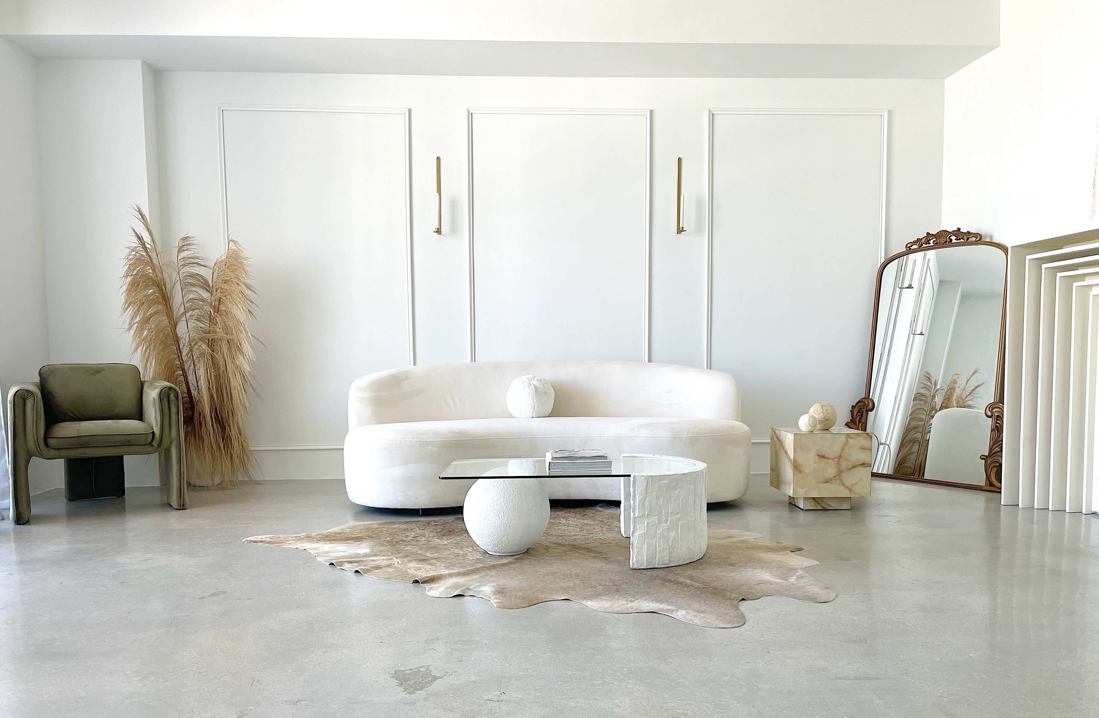 The scene features a minimalist living room with a white curved sofa, a small round coffee table, a decorative mirror, and pampas grass accent decor.