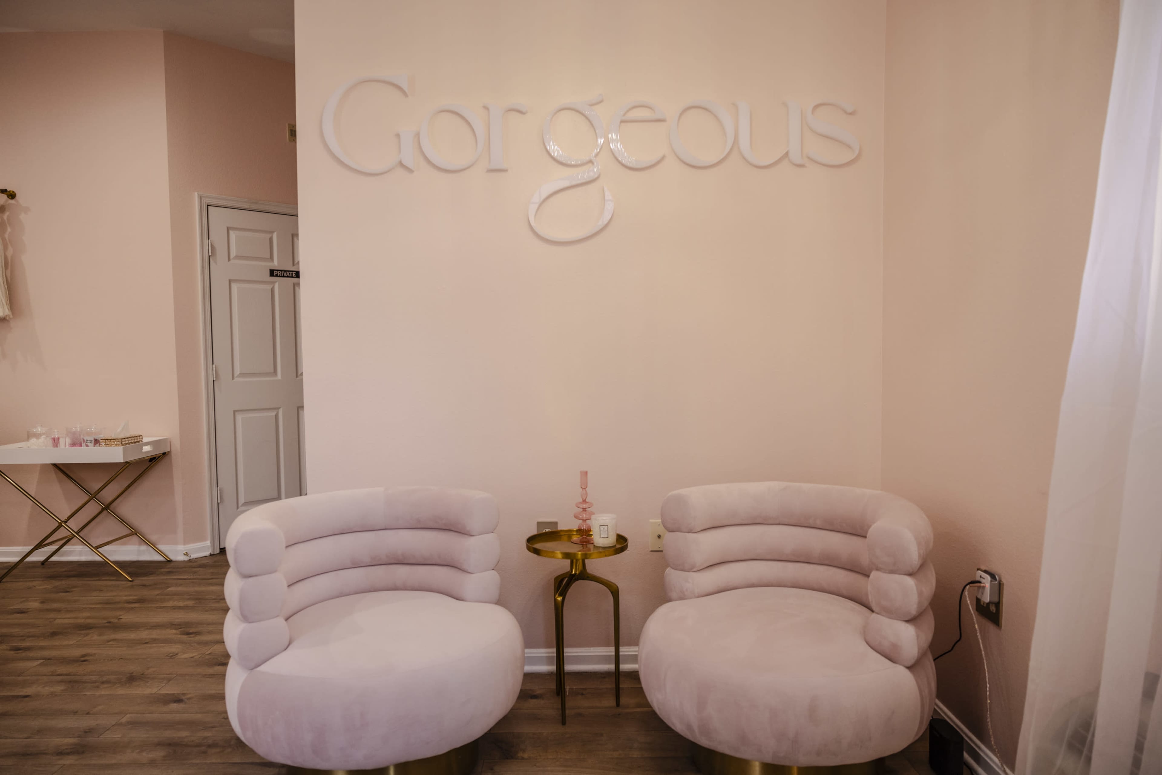 A cozy seating area with two light pink chairs and a small gold side table against a soft pink wall featuring the word "Gorgeous" in large, white letters.