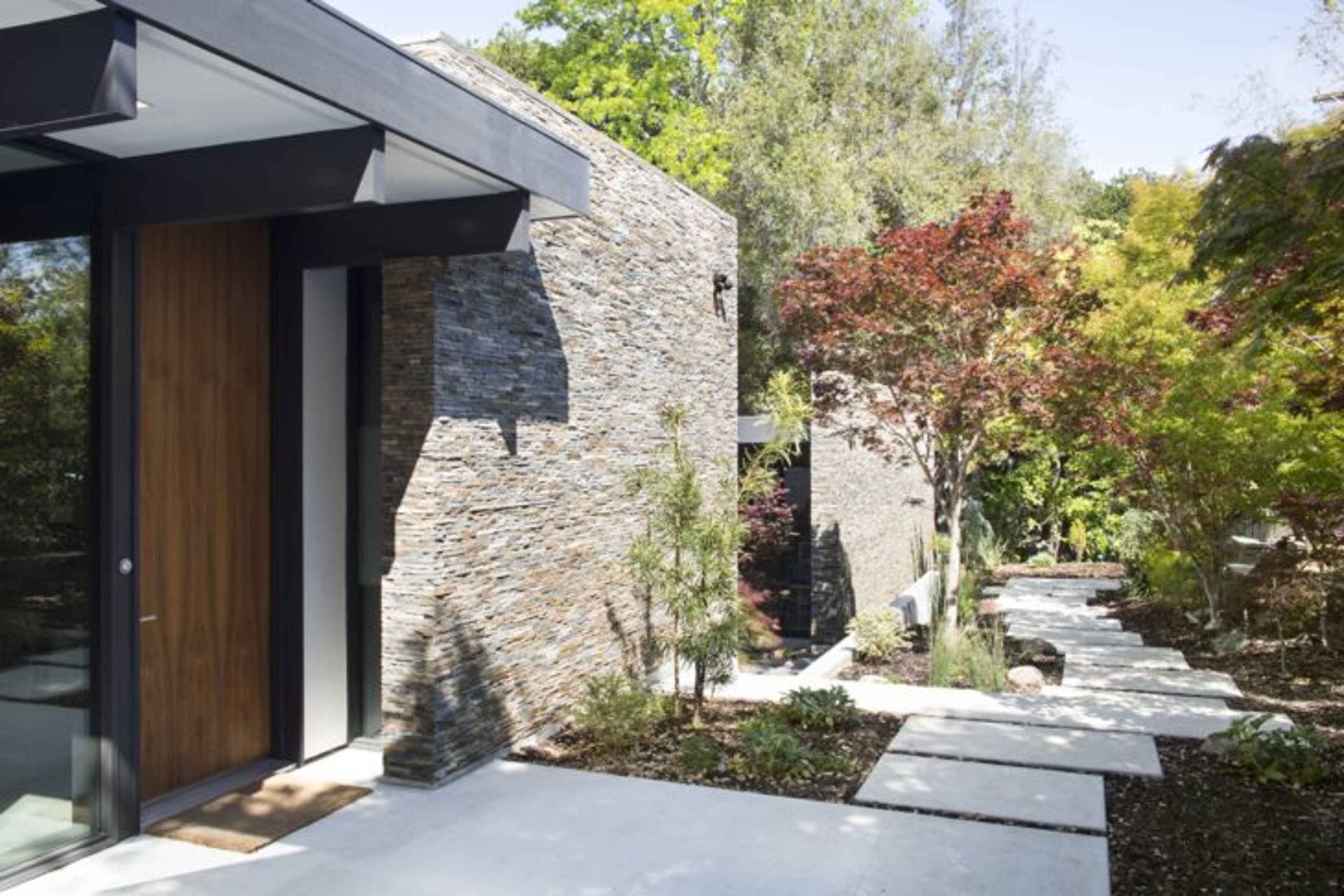 A modern house with stone walls and a concrete pathway leading through a landscaped area.