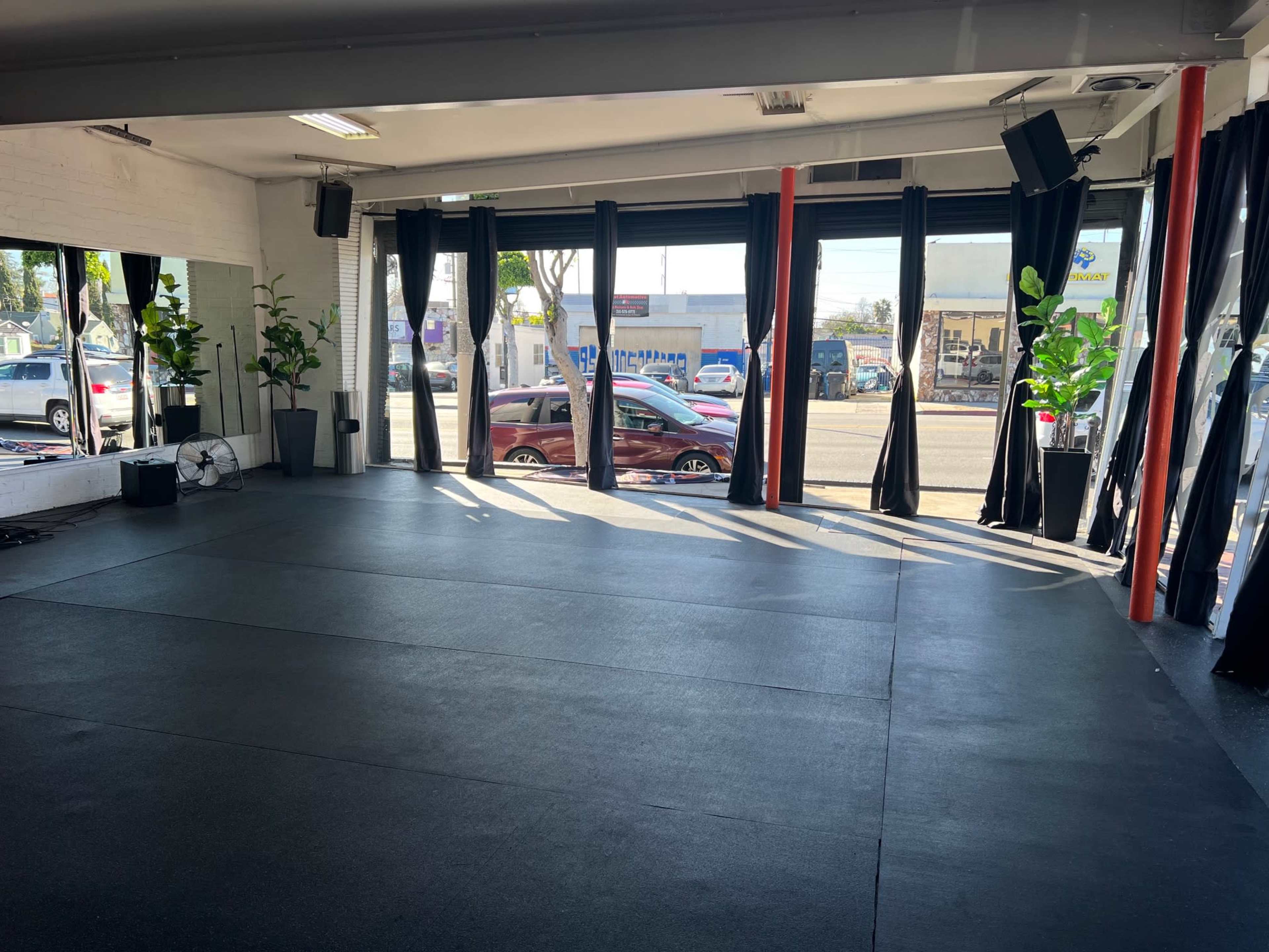 The image shows an empty fitness studio with large windows covered by black curtains, and potted plants in the corners.