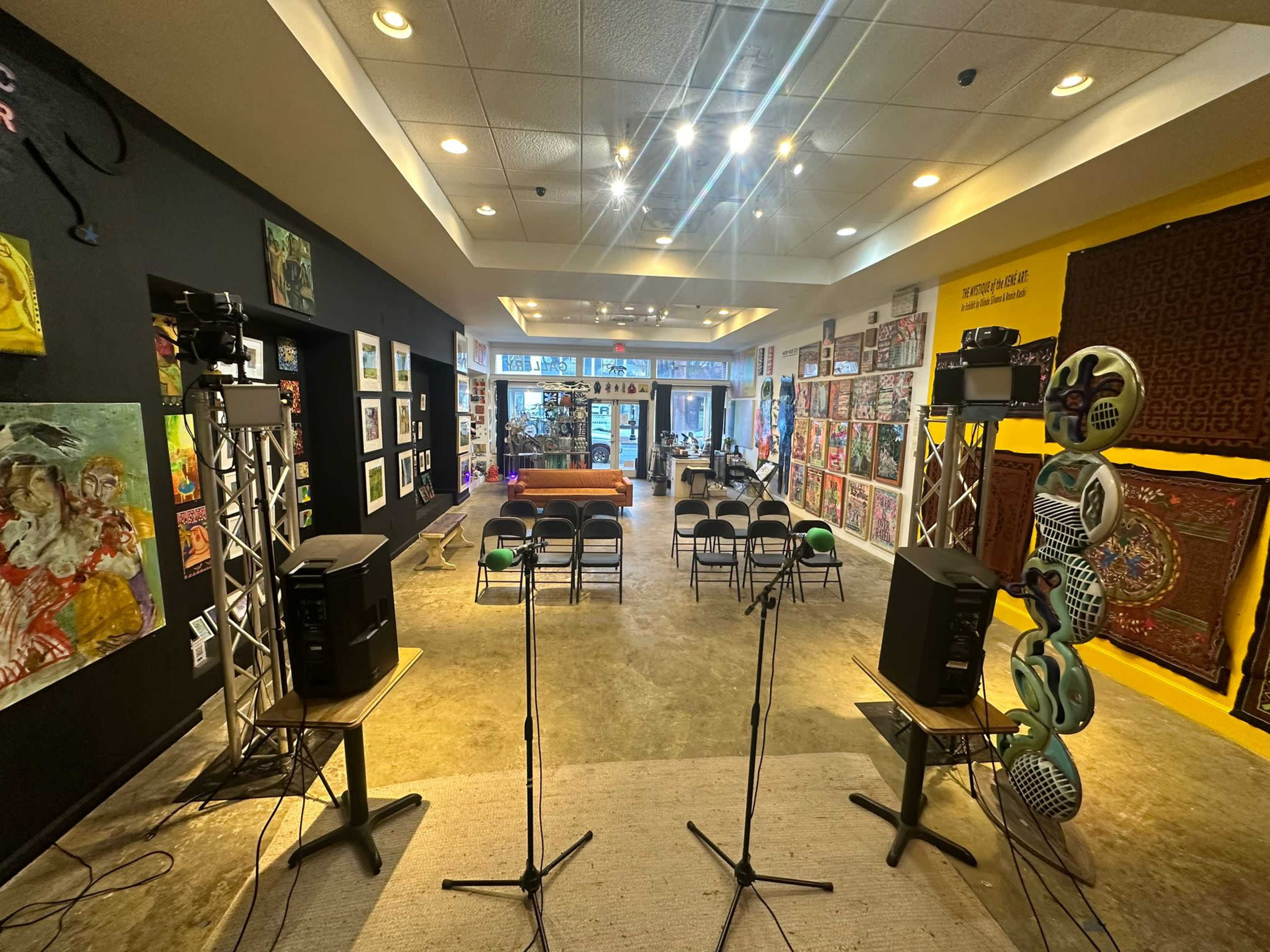 A spacious art gallery with colorful wall-mounted artworks, audio equipment set up on both sides, and rows of chairs arranged for an event.
