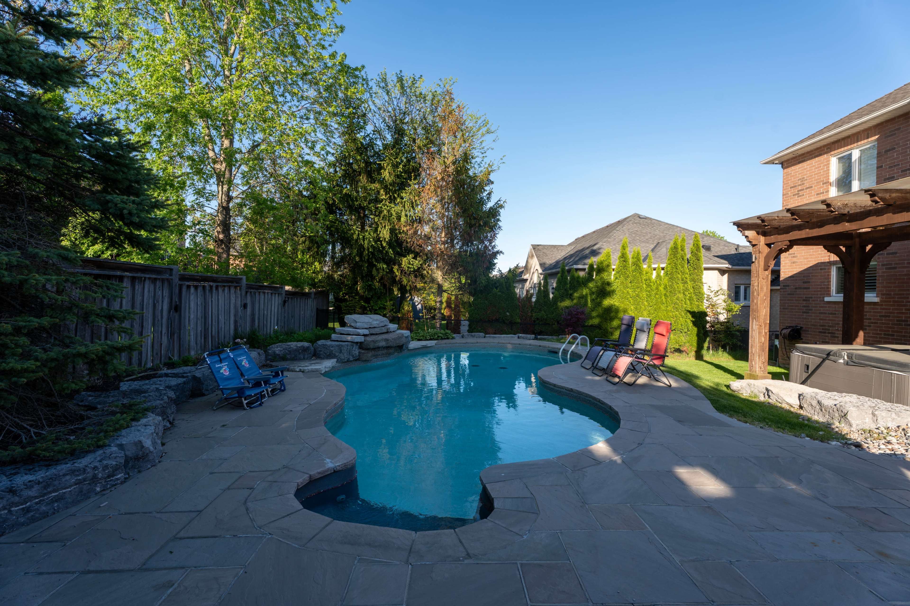 The image shows a residential backyard featuring a swimming pool surrounded by stone decking, chairs, and lush greenery.