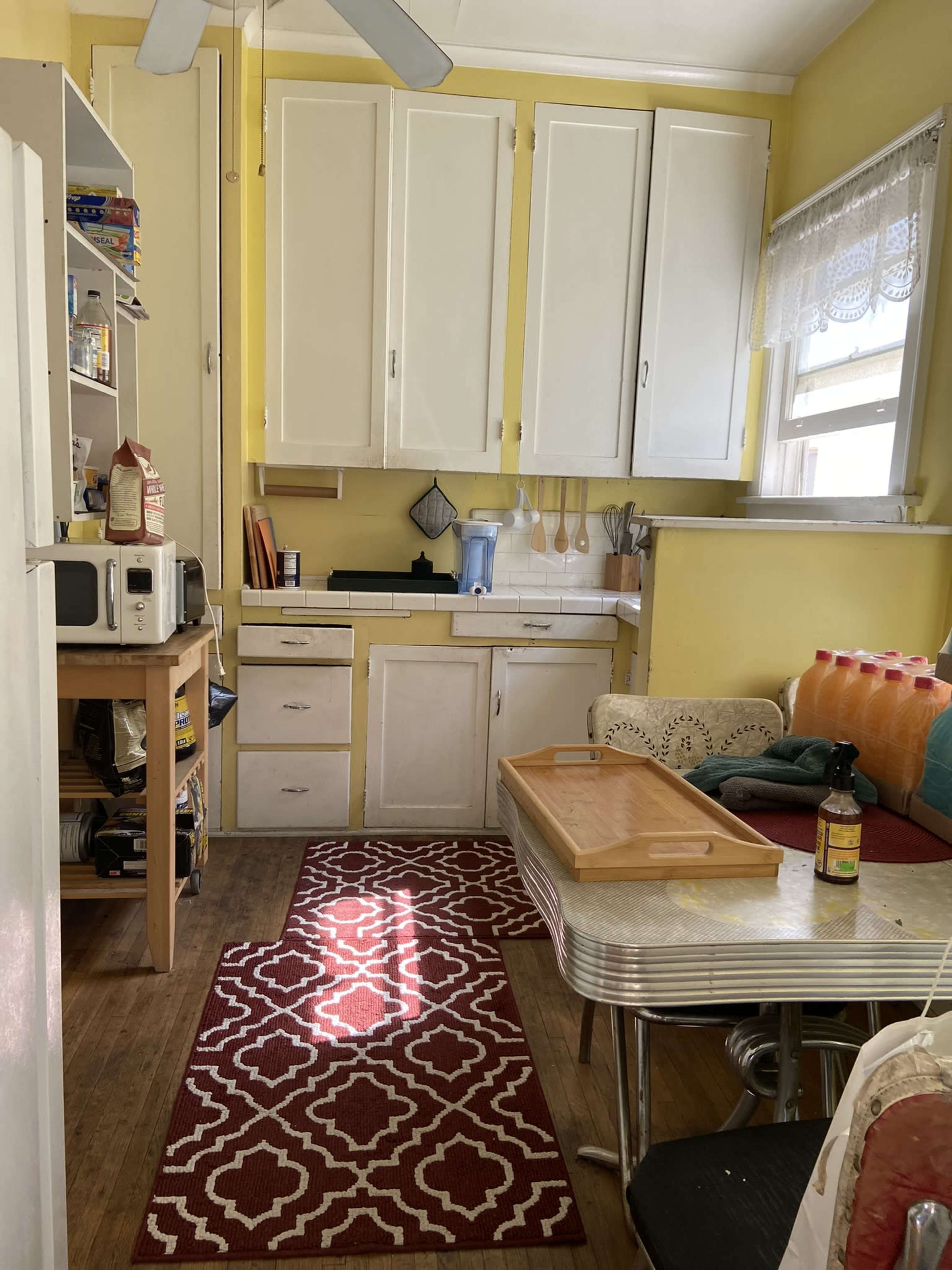 A small kitchen features yellow walls, a red patterned rug, a table with a tray, and wooden cabinets with various kitchen items visible.