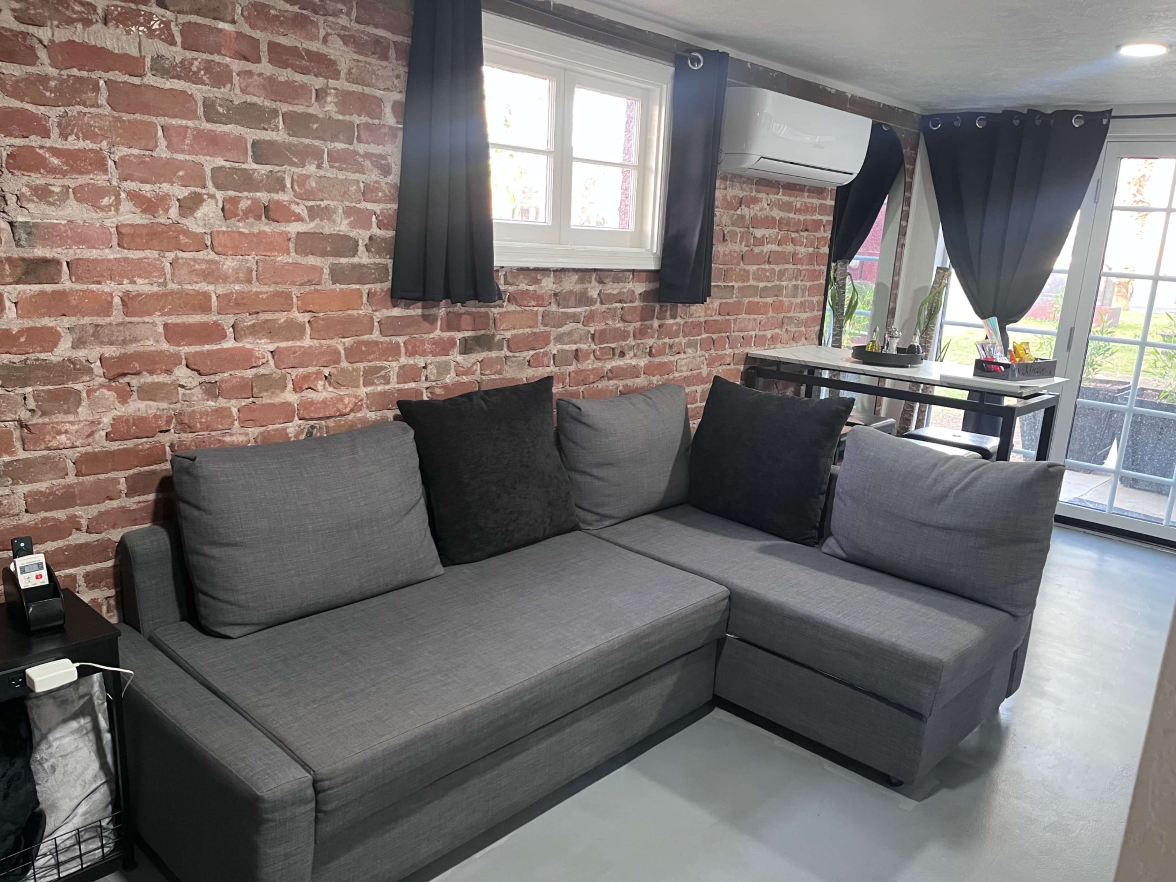 The image shows a living room with a gray sectional couch against a brick wall, accompanied by black curtains and a small dining table near a window.