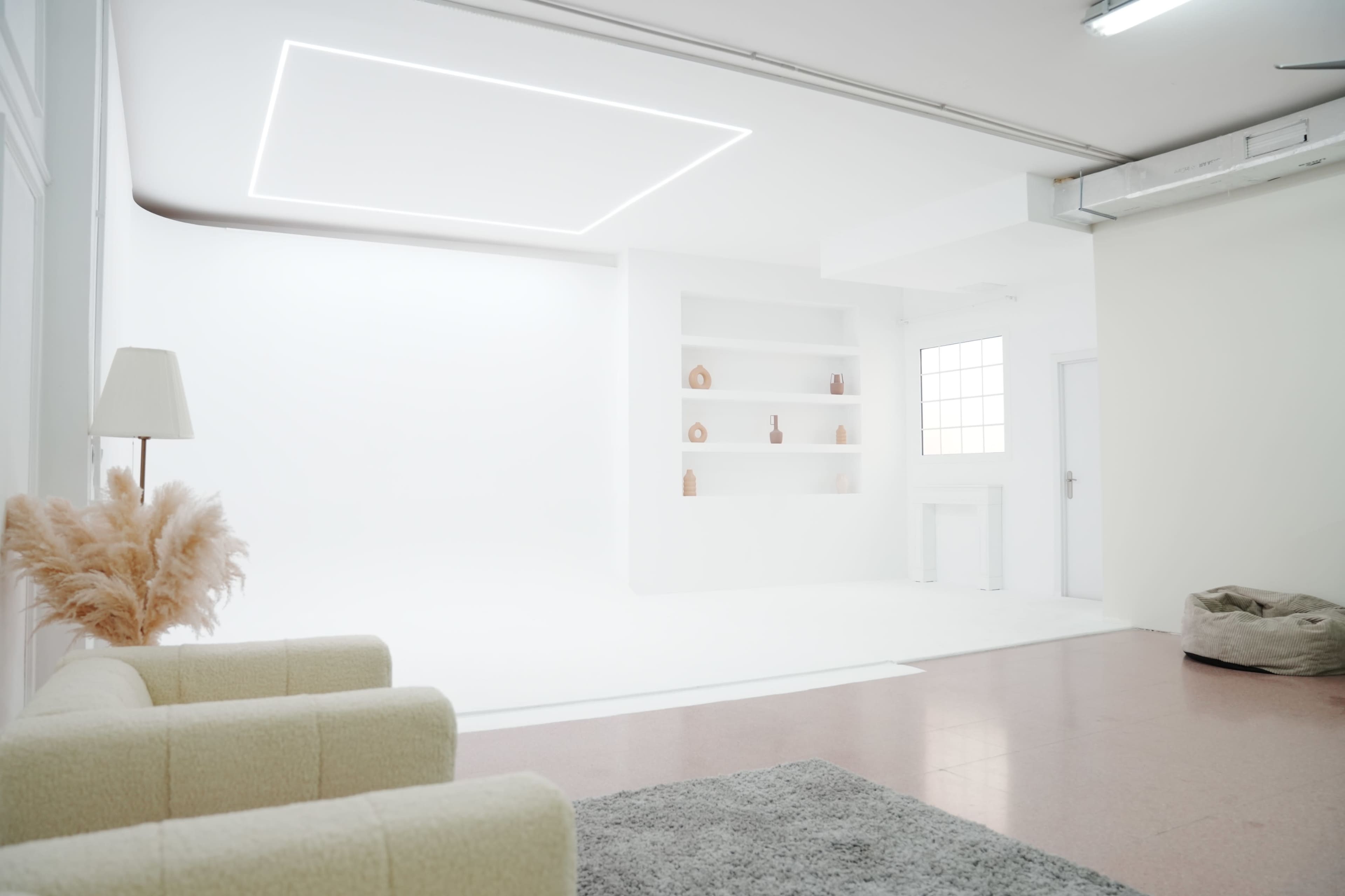 The image showcases a minimalist studio space featuring a light-colored couch, a rug, and a shelf displaying decorative vases against a white backdrop.