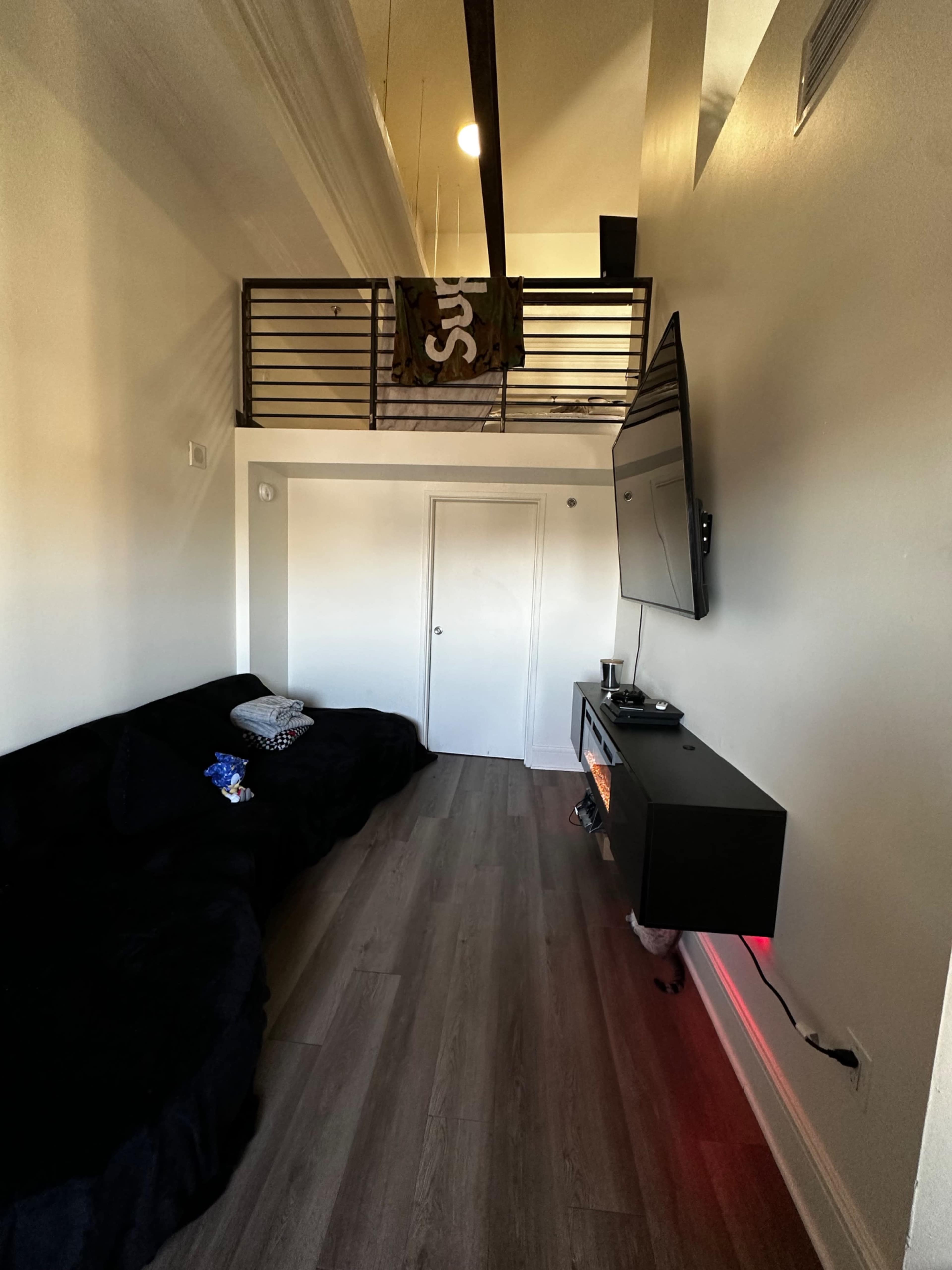 The image shows a modern living space with a dark couch, a wall-mounted television, and a balcony above with a flag hanging.