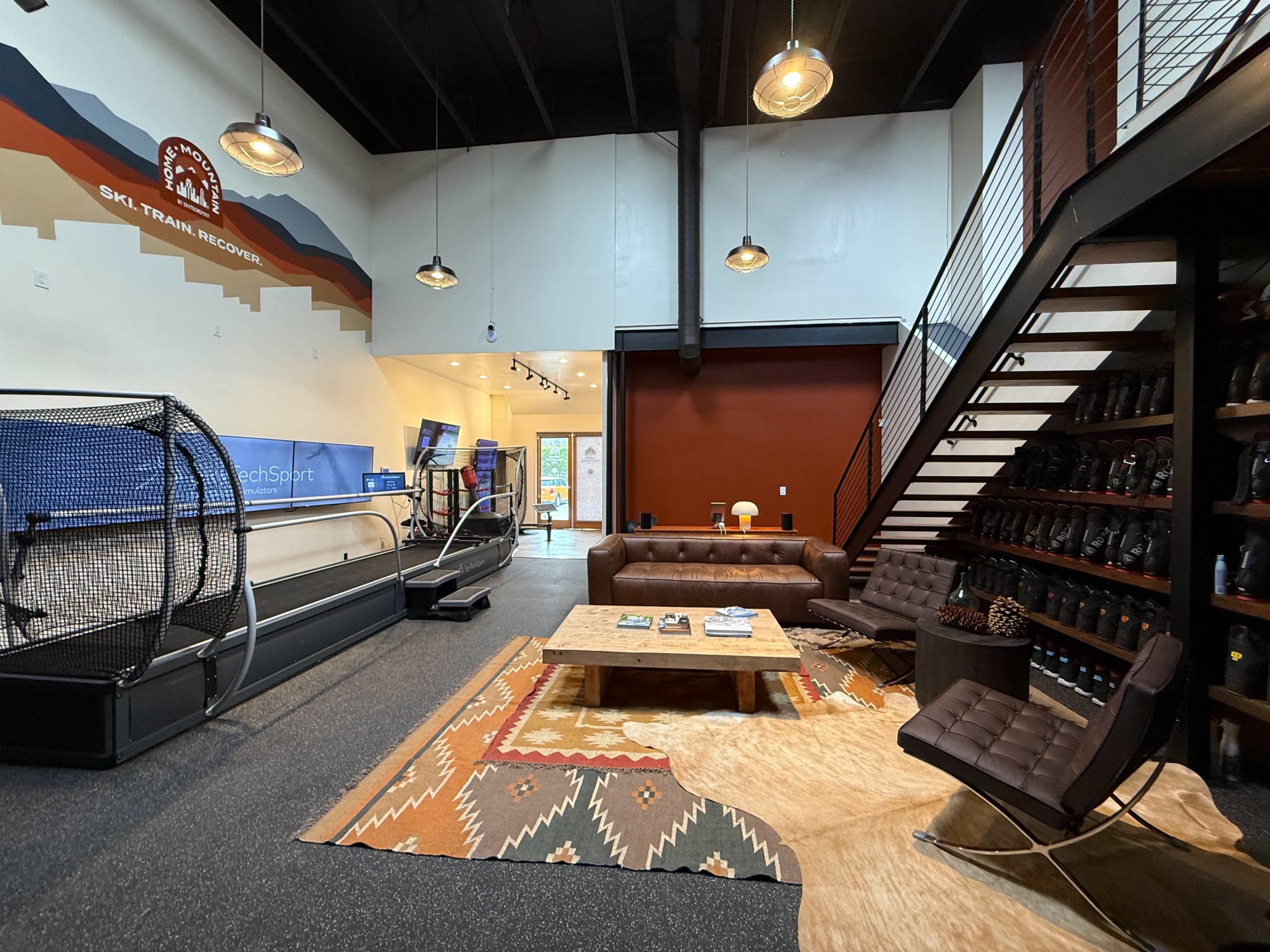 The image shows a spacious interior of a ski resort training facility featuring a treadmill, seating area with brown leather sofas, and a staircase leading to an upper level.