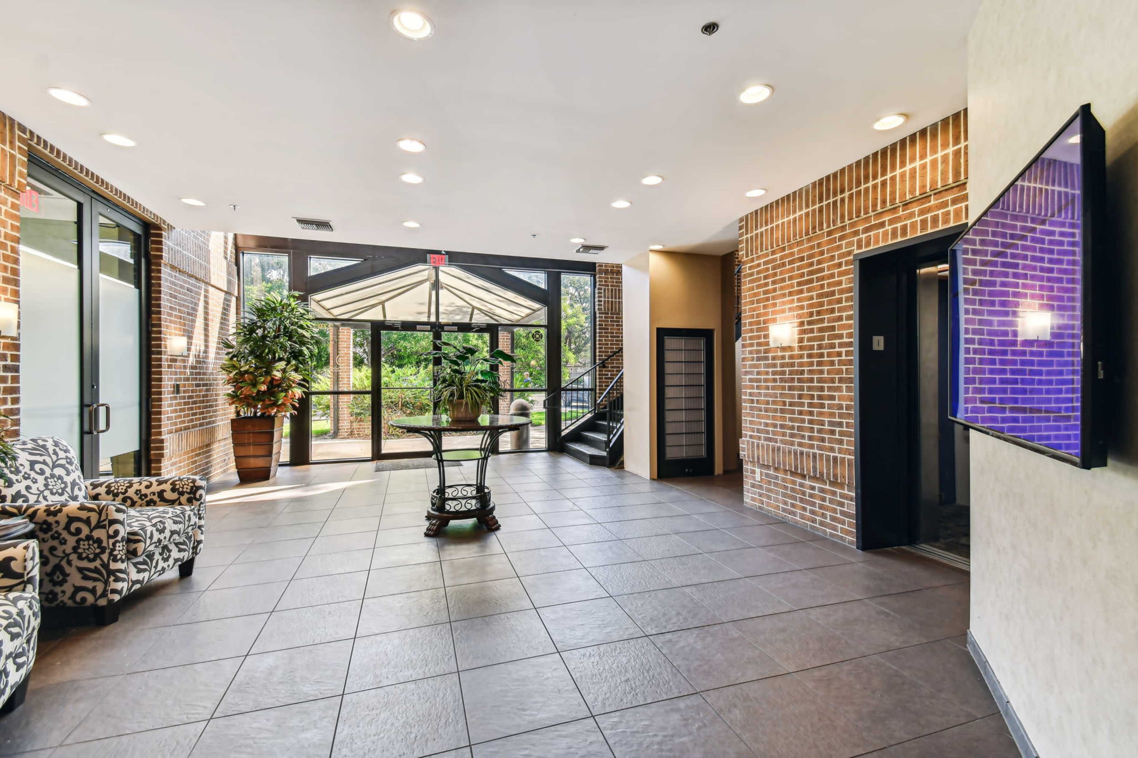The image shows a modern lobby area with large windows, brick walls, tiled flooring, and a seating area featuring patterned chairs and a decorative plant.