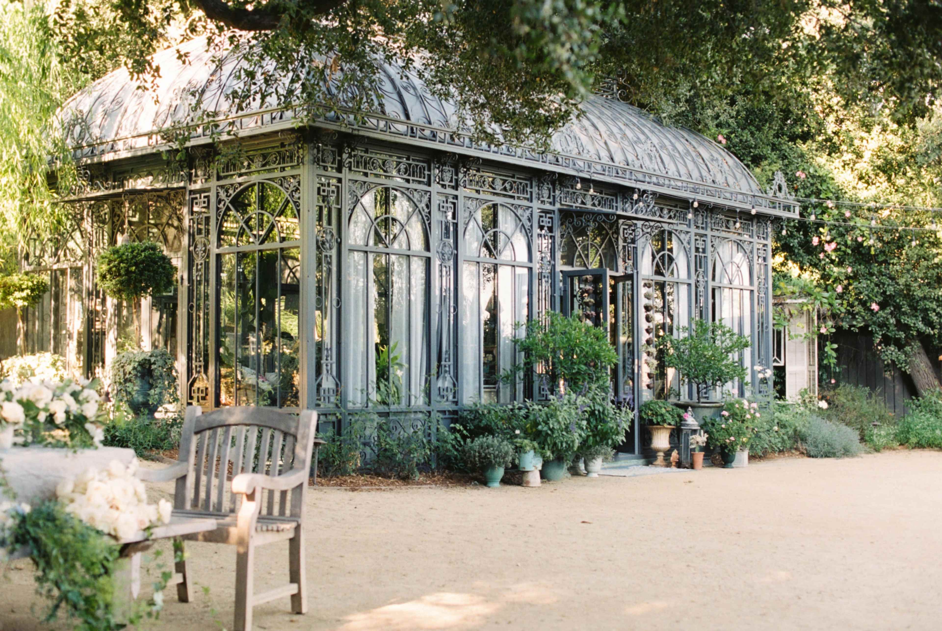 A glass conservatory with intricate metalwork is surrounded by greenery and potted plants in a garden setting.
