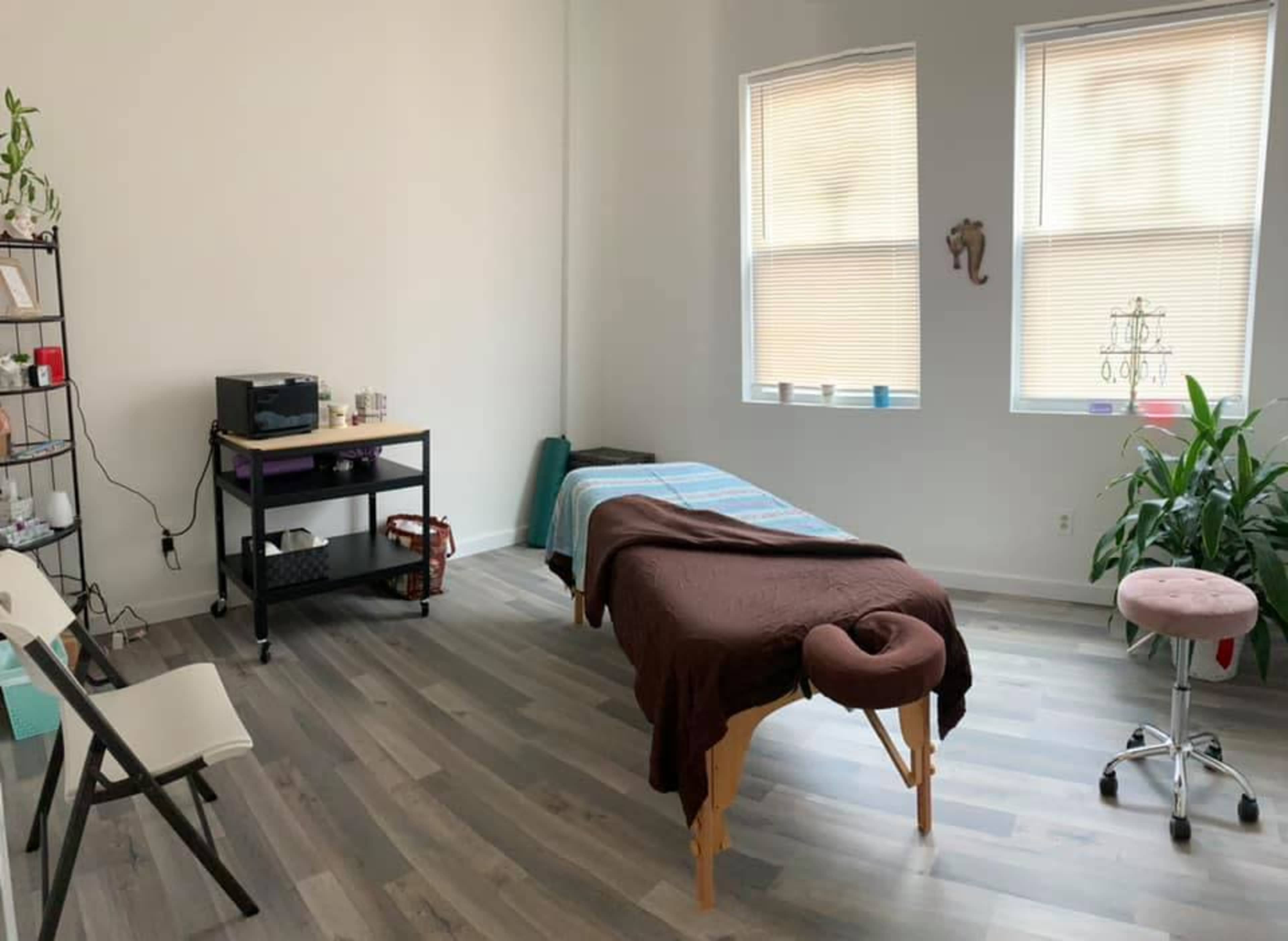The image shows a massage room with a treatment table, seating, and natural light coming through the windows.