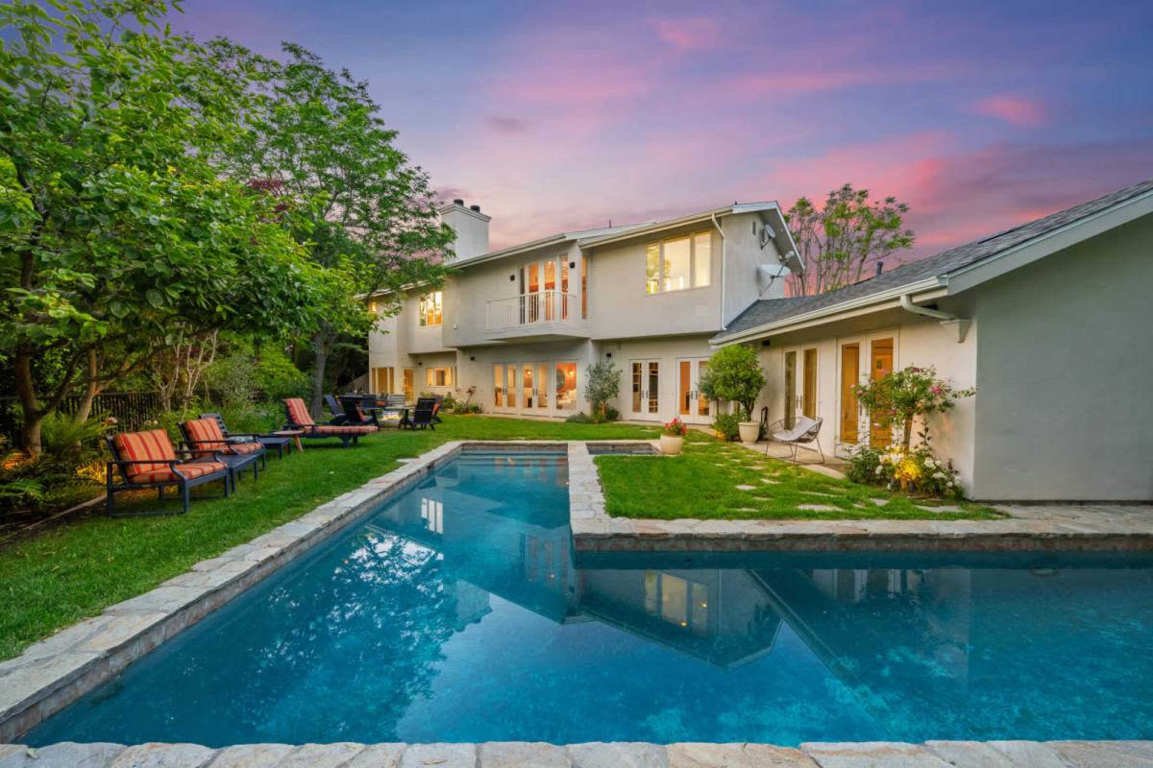 A spacious backyard features a swimming pool surrounded by lush greenery and modern lounge chairs near a two-story house at sunset.