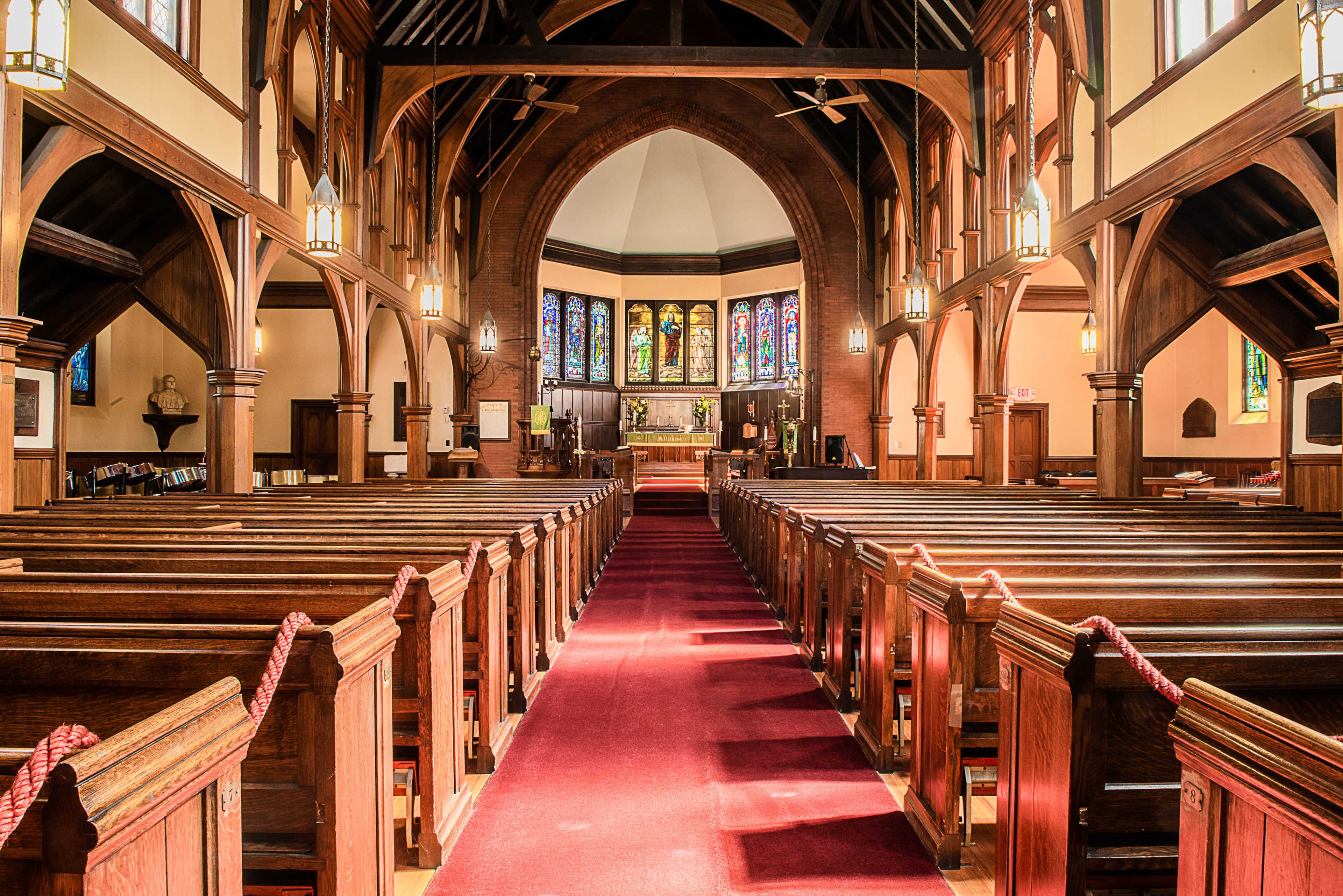 The interior of a wooden church features neatly arranged pews, a red aisle runner, and stained glass windows illuminating the altar at the front.