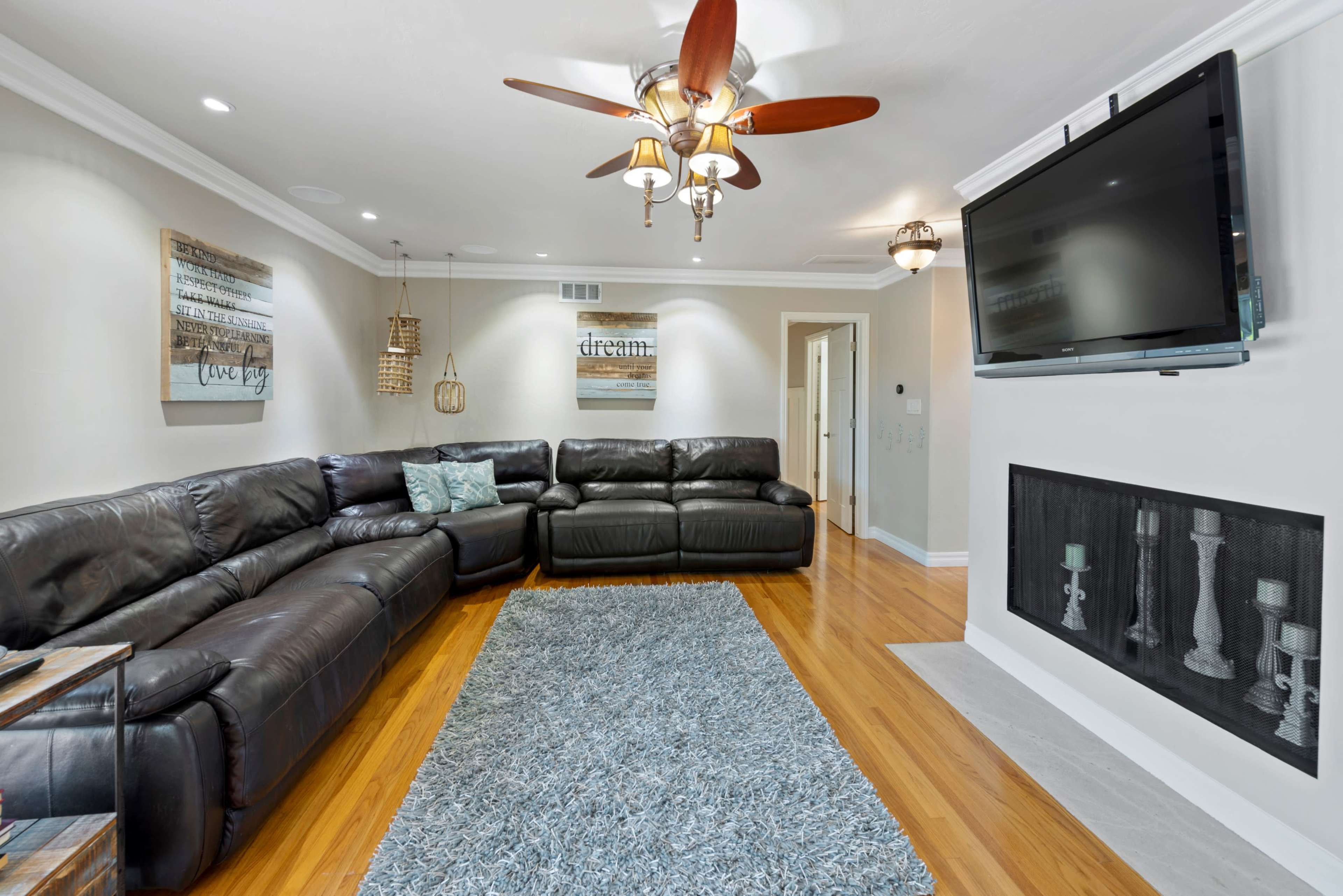 The living room features a large leather sectional sofa, a flat-screen TV mounted on the wall, and a decorative area rug on polished hardwood floors.