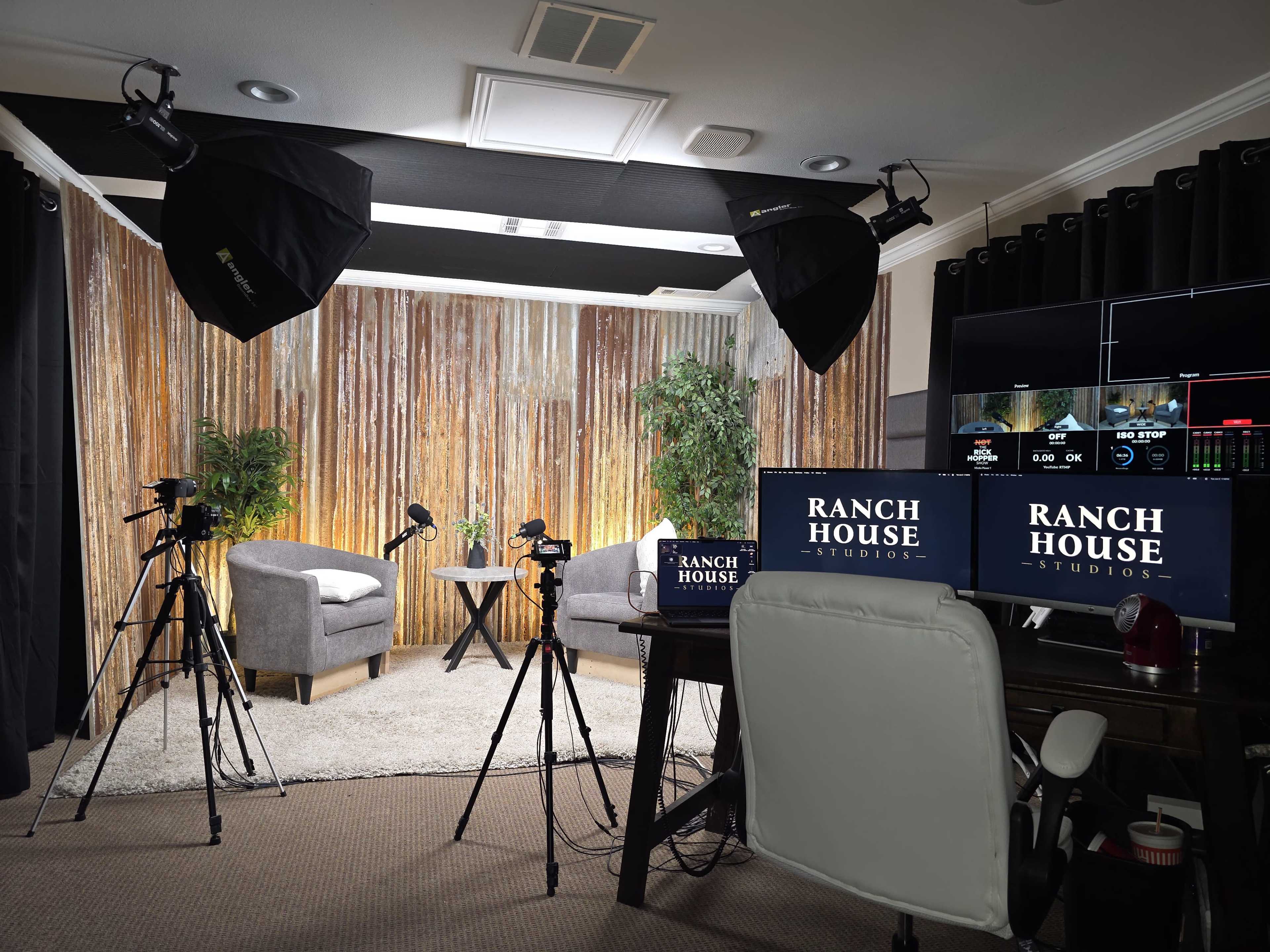 The image shows a studio setup with two gray chairs, a small table, lighting equipment, and multiple monitors displaying a control screen, all arranged against a backdrop of decorative curtains.