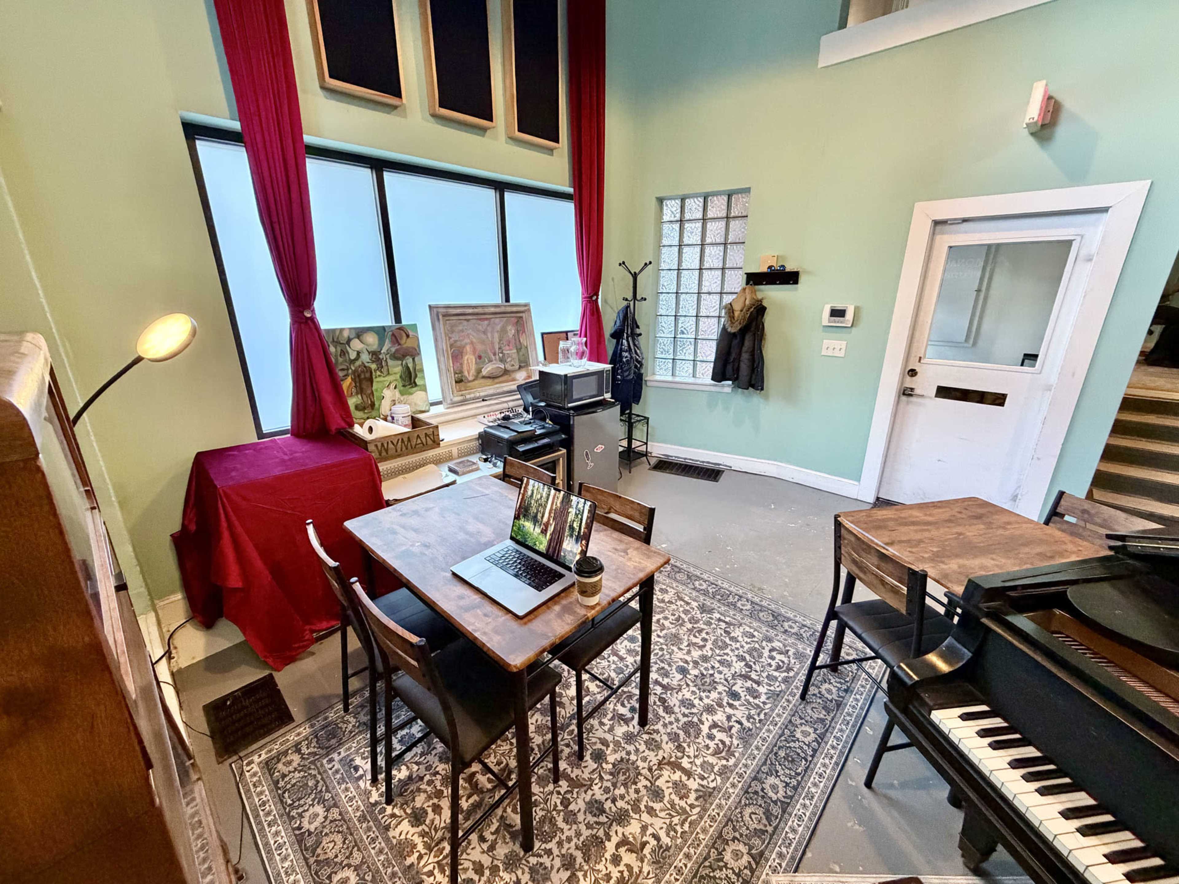The image shows a spacious room with a table, chairs, and a laptop, along with a piano and large windows featuring red curtains.