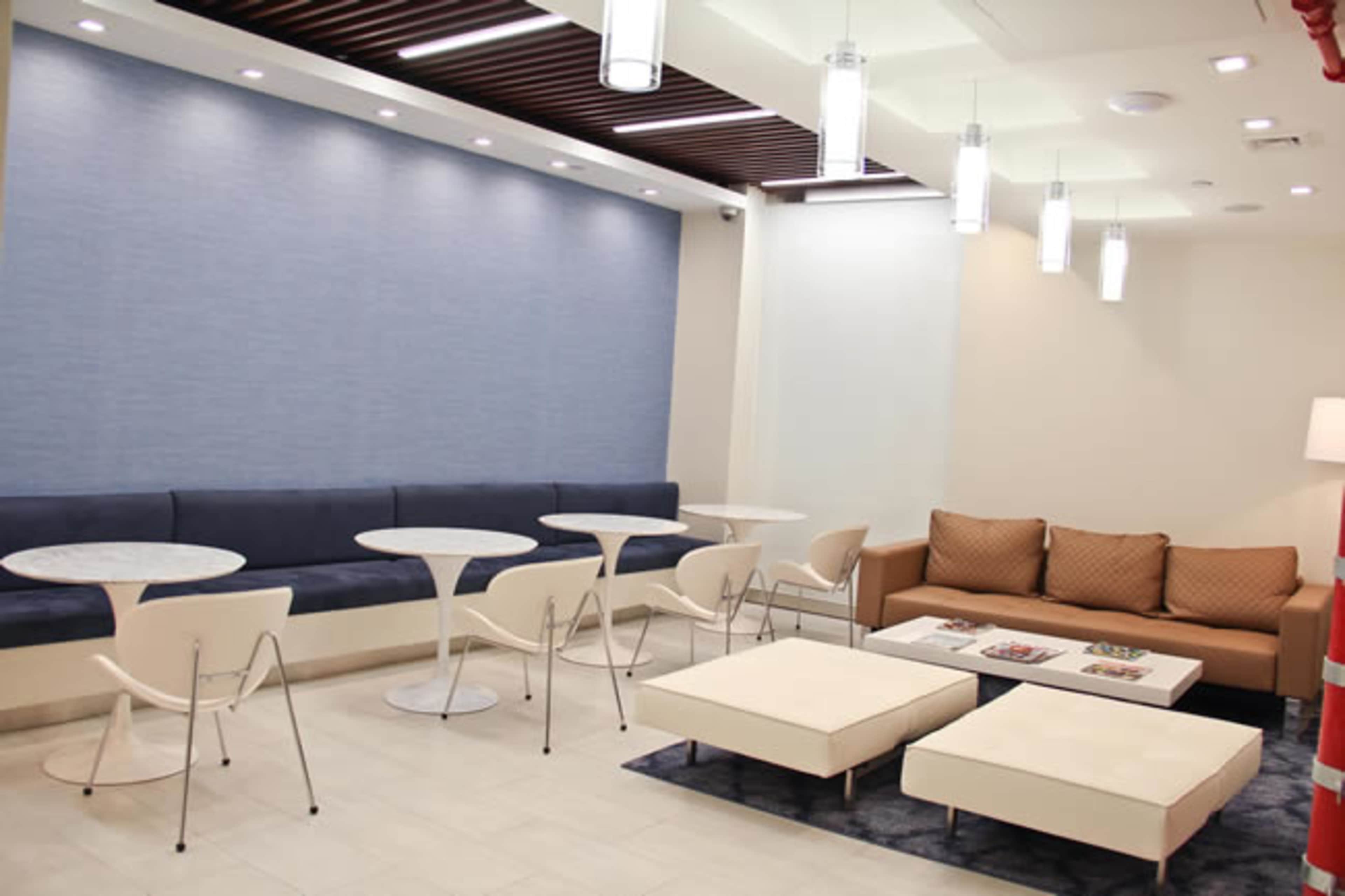 A modern lounge area with a blue accent wall, white tables, and seating arrangements including a beige couch and plush ottomans.