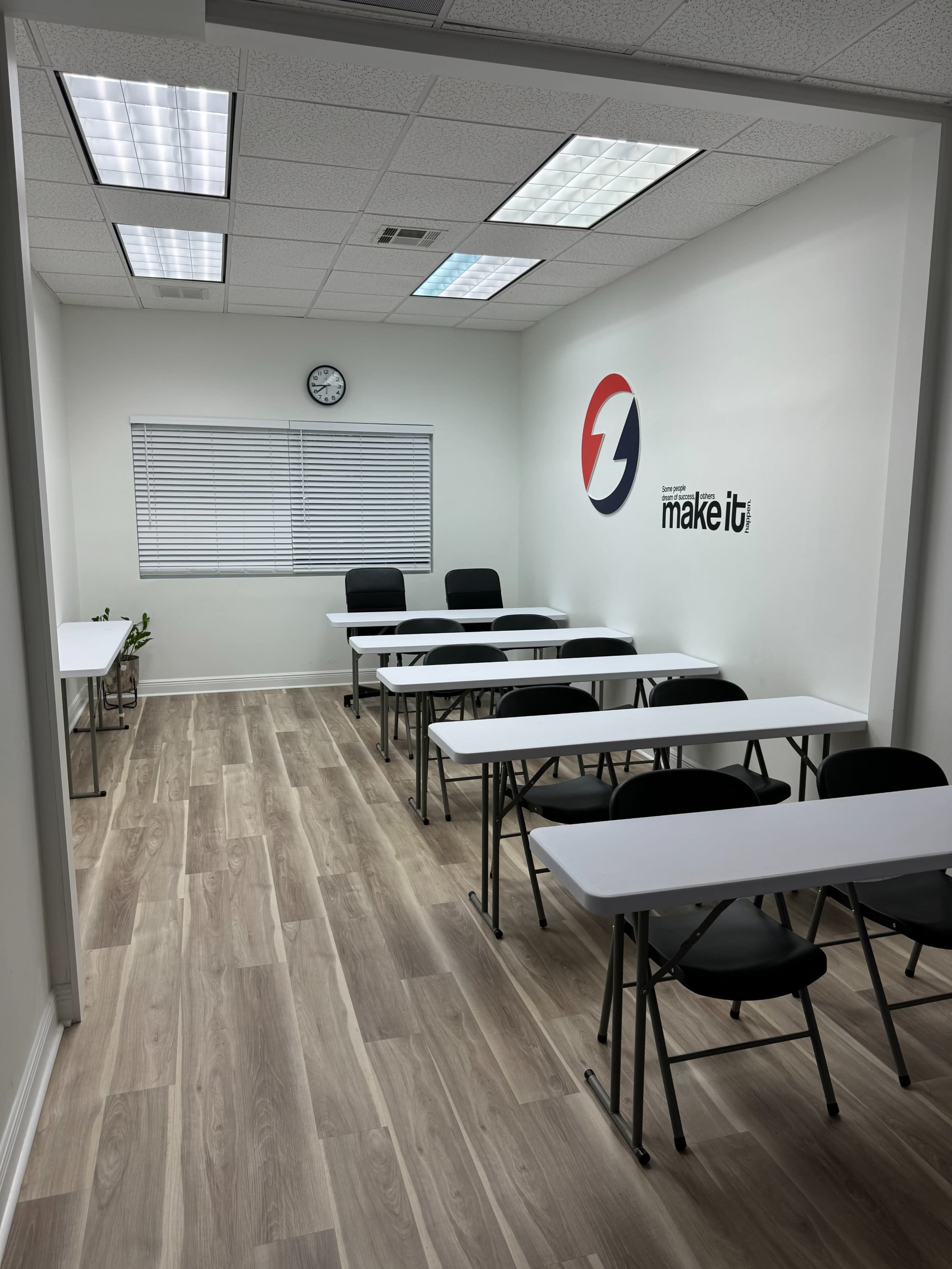 The image shows a classroom with several white tables and black chairs arranged neatly, along with a wall clock and a logo on the wall.