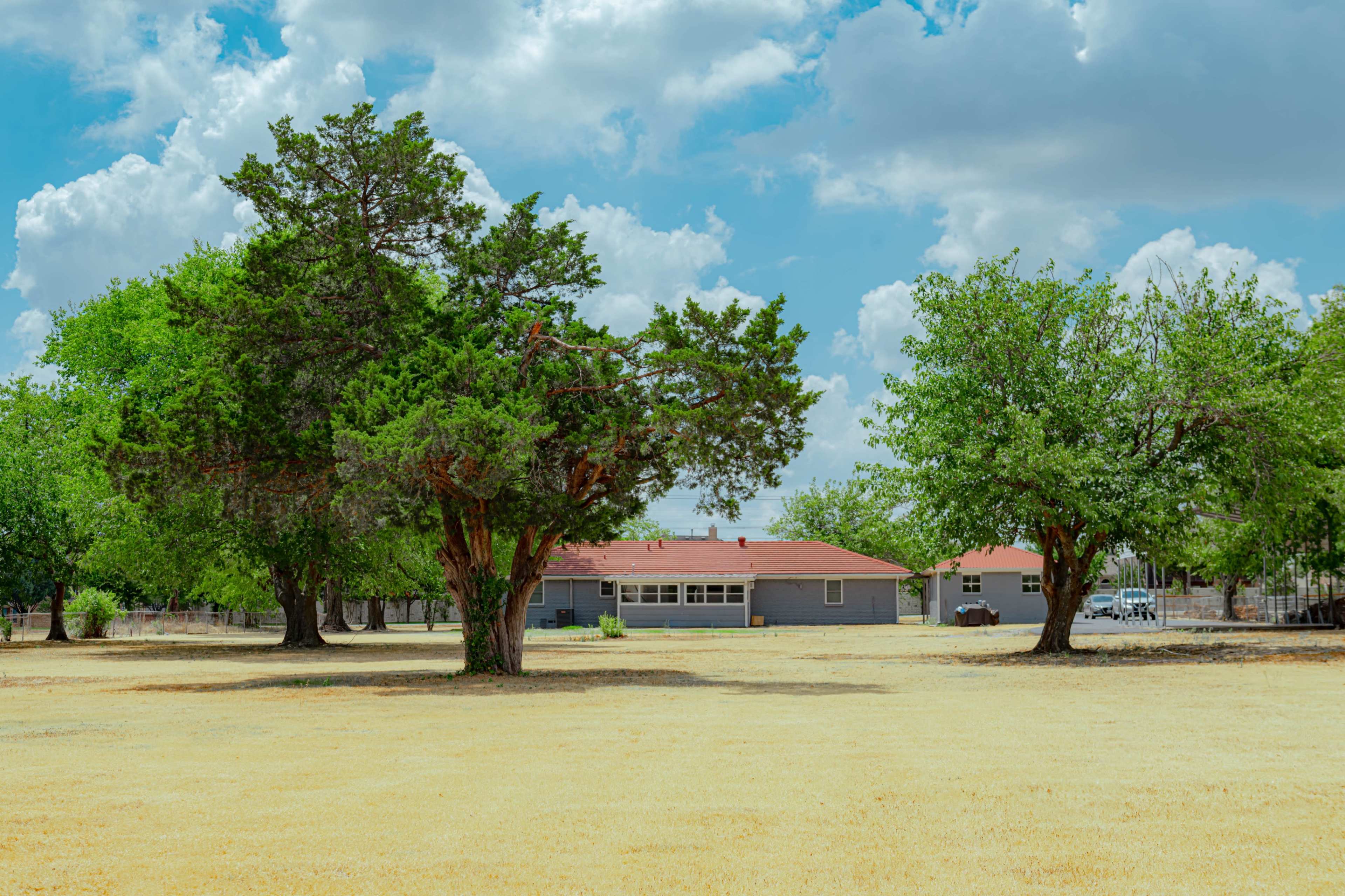 A gray house with a red roof is set in a grassy field surrounded by several green trees under a partly cloudy sky.