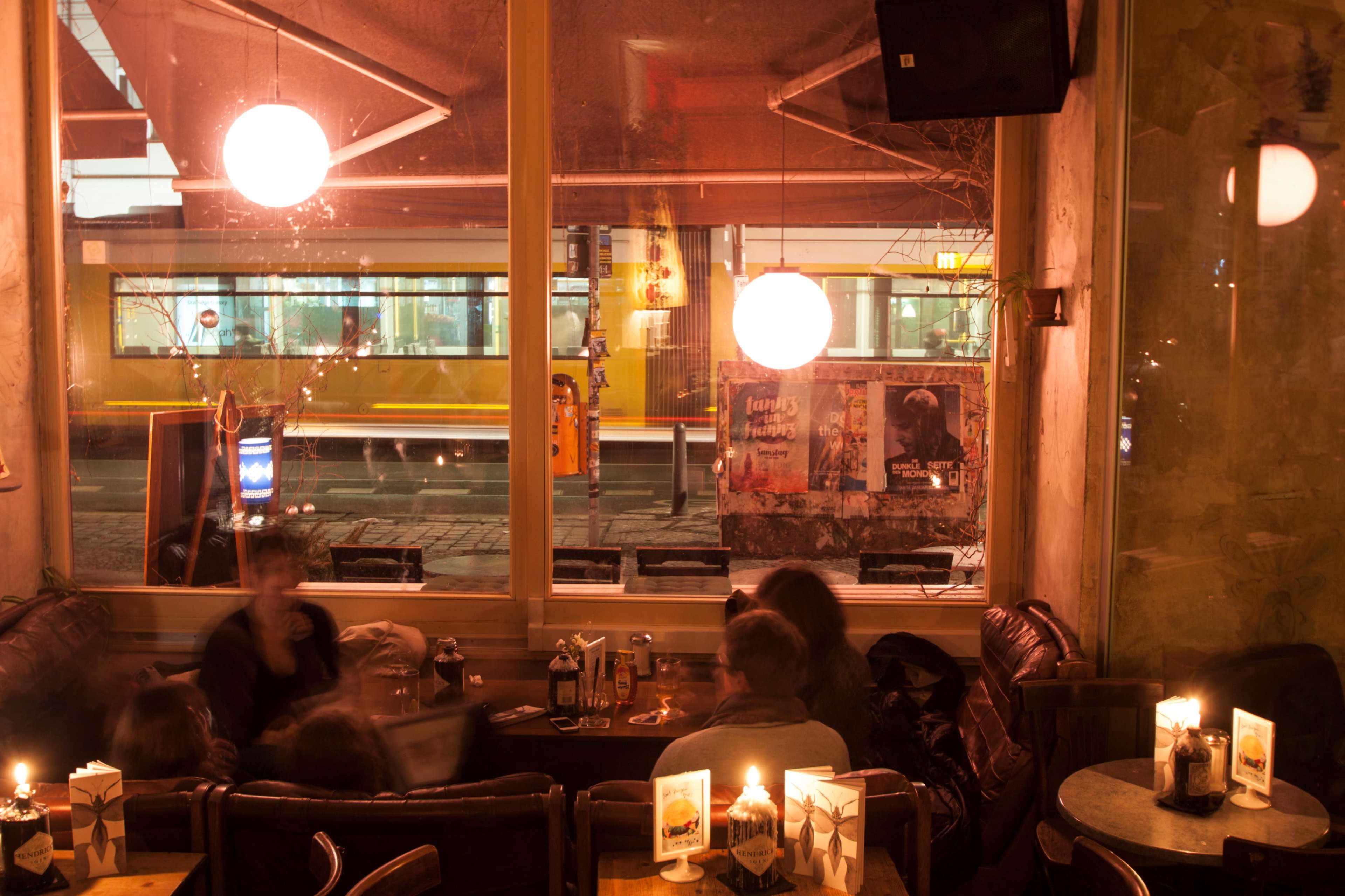 A cozy café scene is illuminated by candles, with patrons seated at tables while a tram passes by outside.