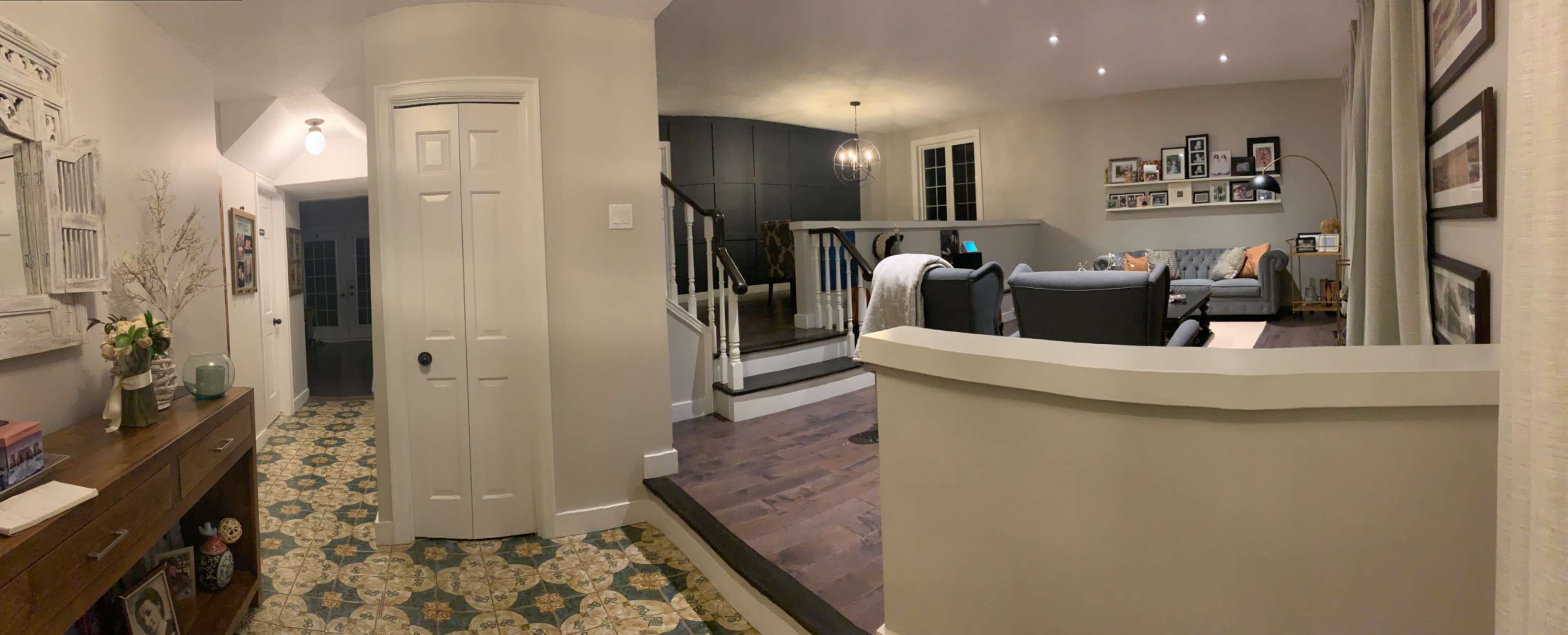 The image shows a spacious interior with a hallway leading to a living area, featuring a sofa, photographs on the walls, and an open staircase.