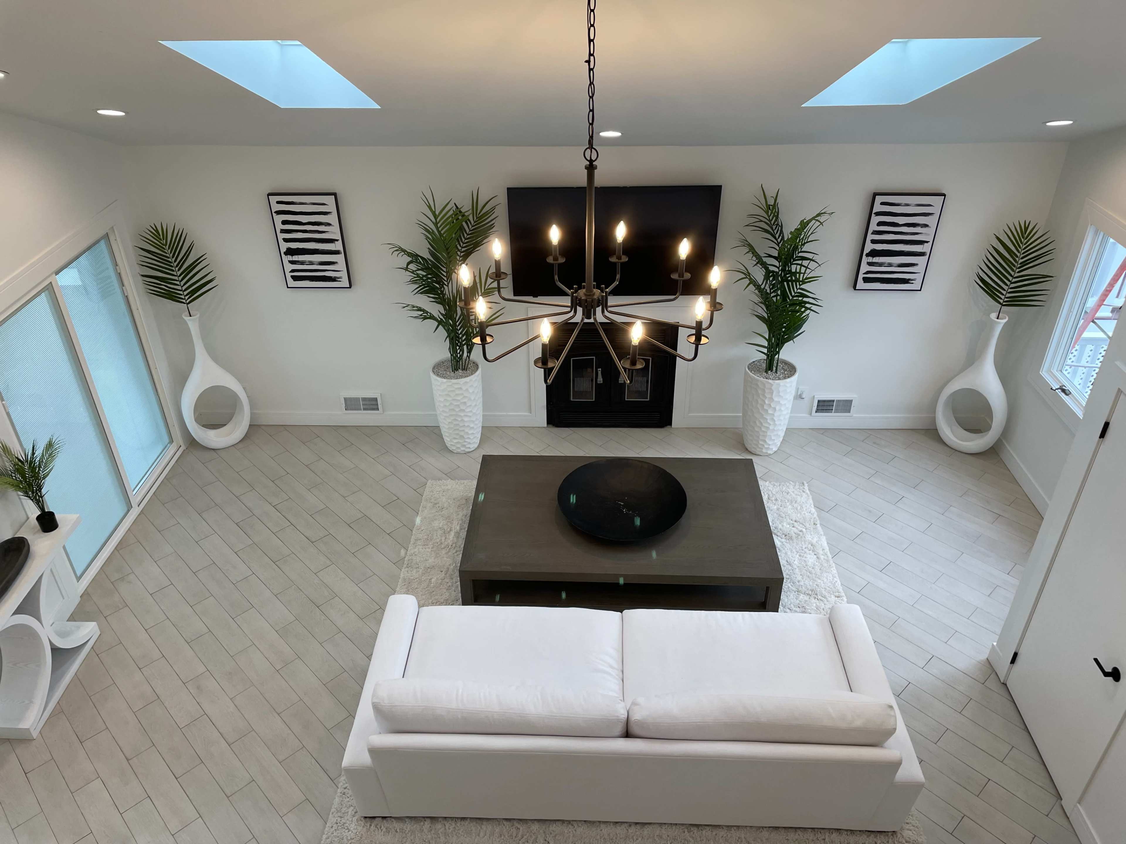 The image shows a modern living room with a white sectional sofa, a dark wooden coffee table, and decorative plants in white planters, centered around a flat-screen TV mounted above a fireplace.