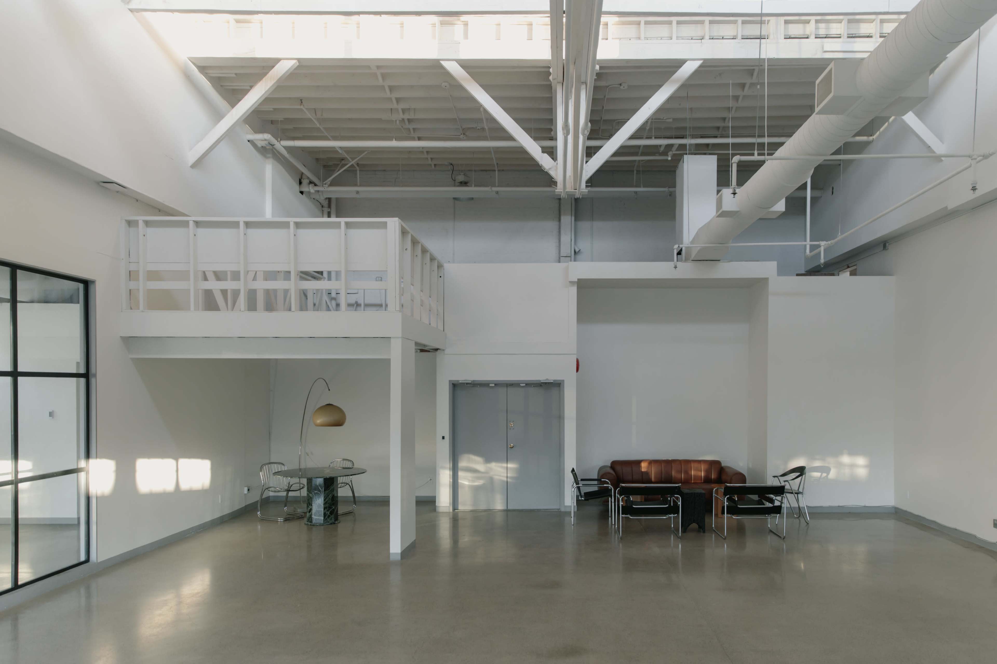 The interior of a spacious, minimalist loft features a mezzanine, a brown leather sofa, a glass dining table with chairs, and large windows allowing natural light to enter.